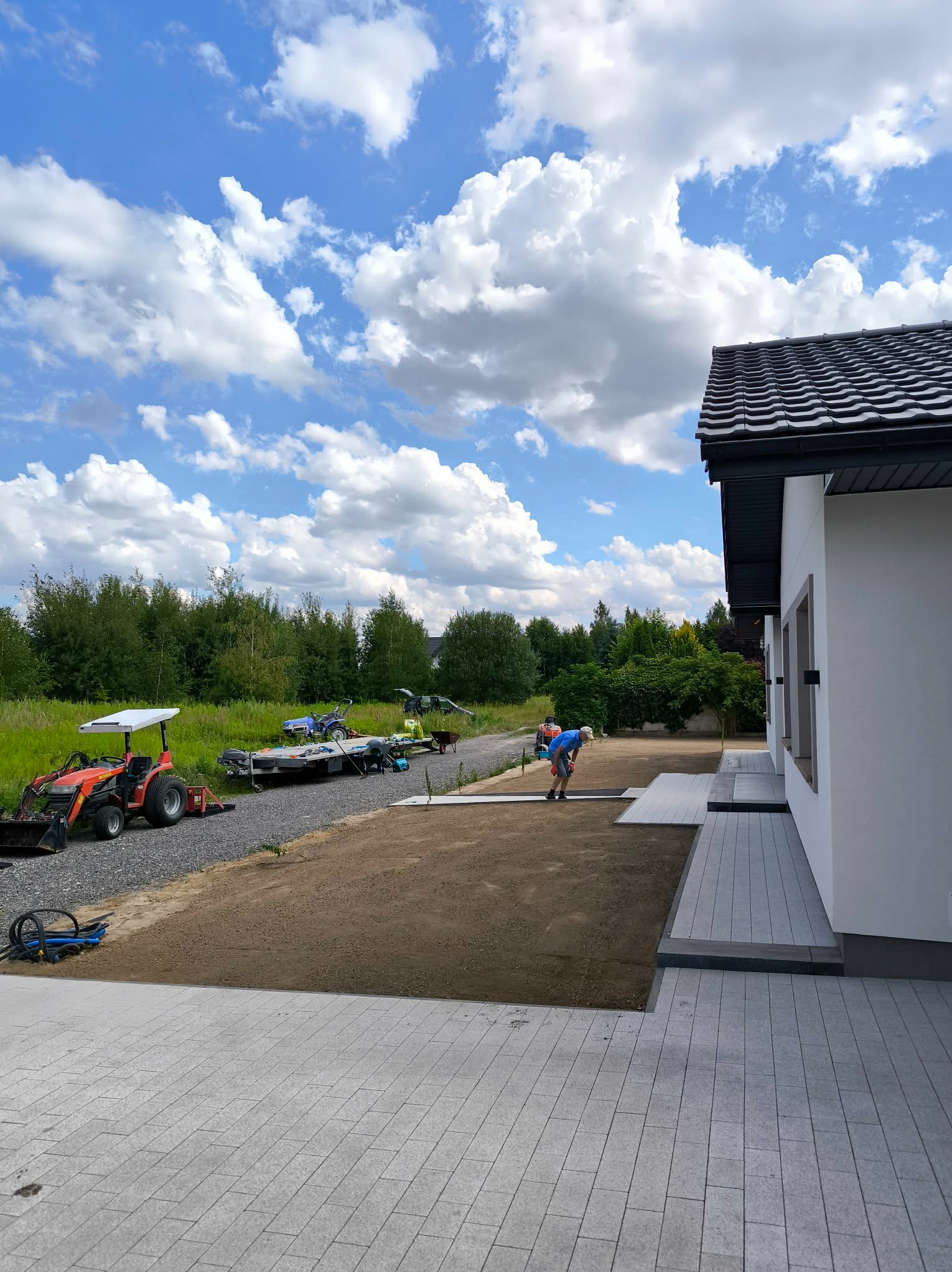 Układanie kostki brukowej na posesji domu jednorodzinnego. Widoczny traktor, przygotowany teren i pracownik w trakcie pracy. Błękitne niebo z chmurami w tle.