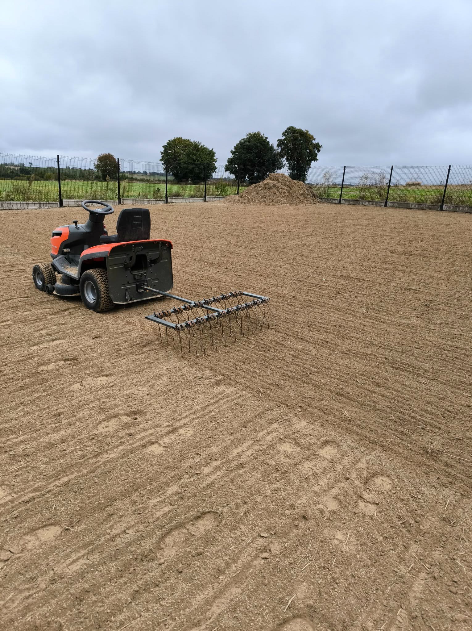 Traktor ogrodowy z broną w Wierzbicy przygotowuje teren pod trawnik. Widoczne ślady pracy na ziemi, ogrodzenie i drzewa w tle. Pochmurne niebo.