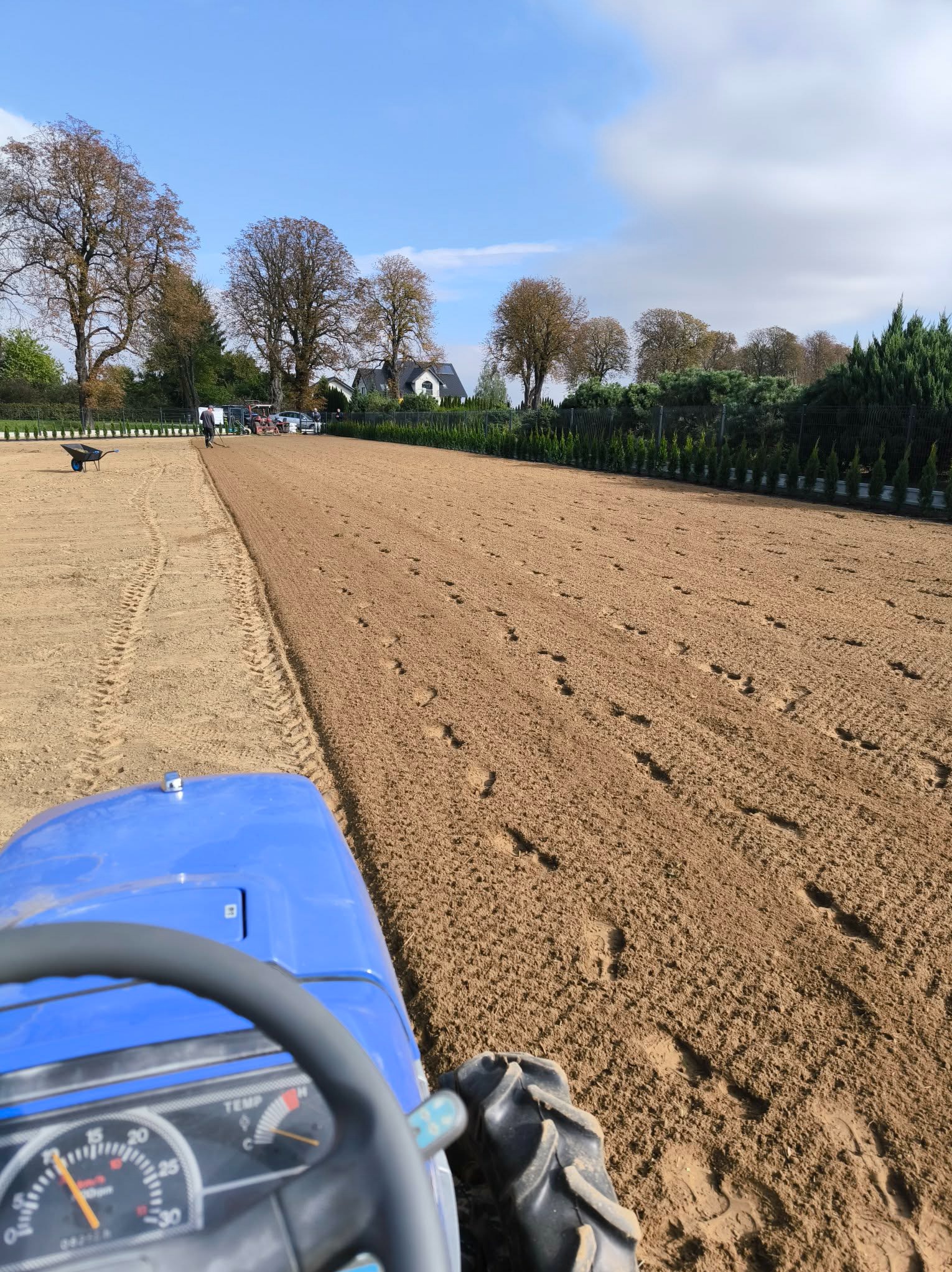 Niebieski traktor przygotowuje teren pod trawnik, widok z perspektywy operatora. W tle drzewa i budynek. Ślady opon na piasku.