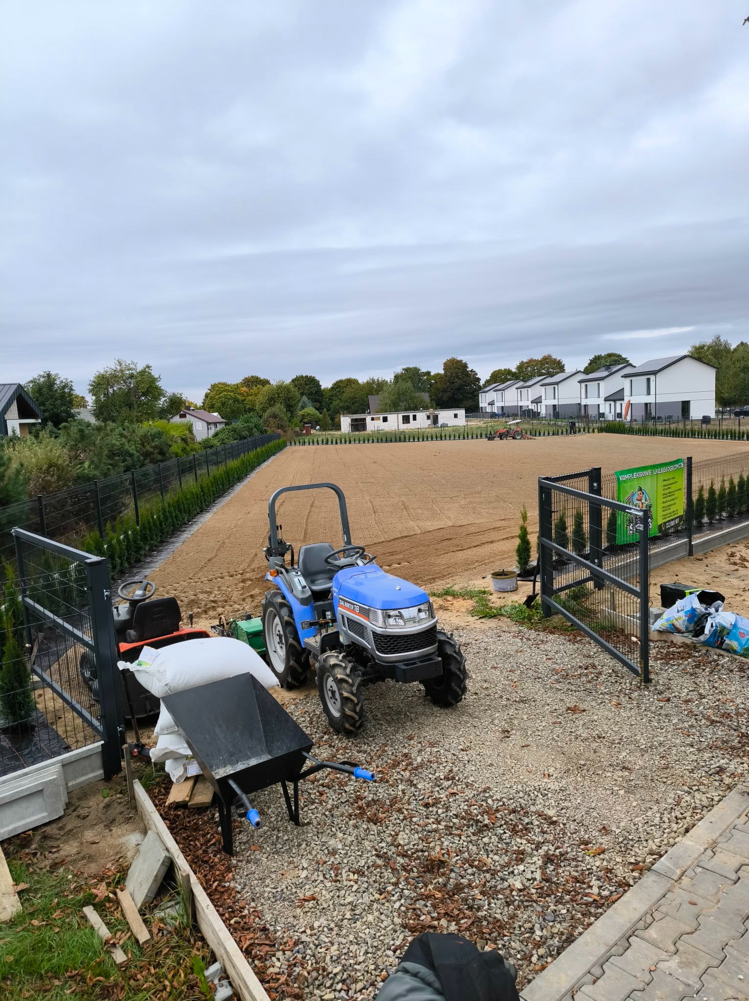 Przygotowany teren pod trawnik z widocznym traktorkiem, taczką i workami. W tle rząd domów i ogrodzenie z nasadzeniami. Pochmurne niebo.