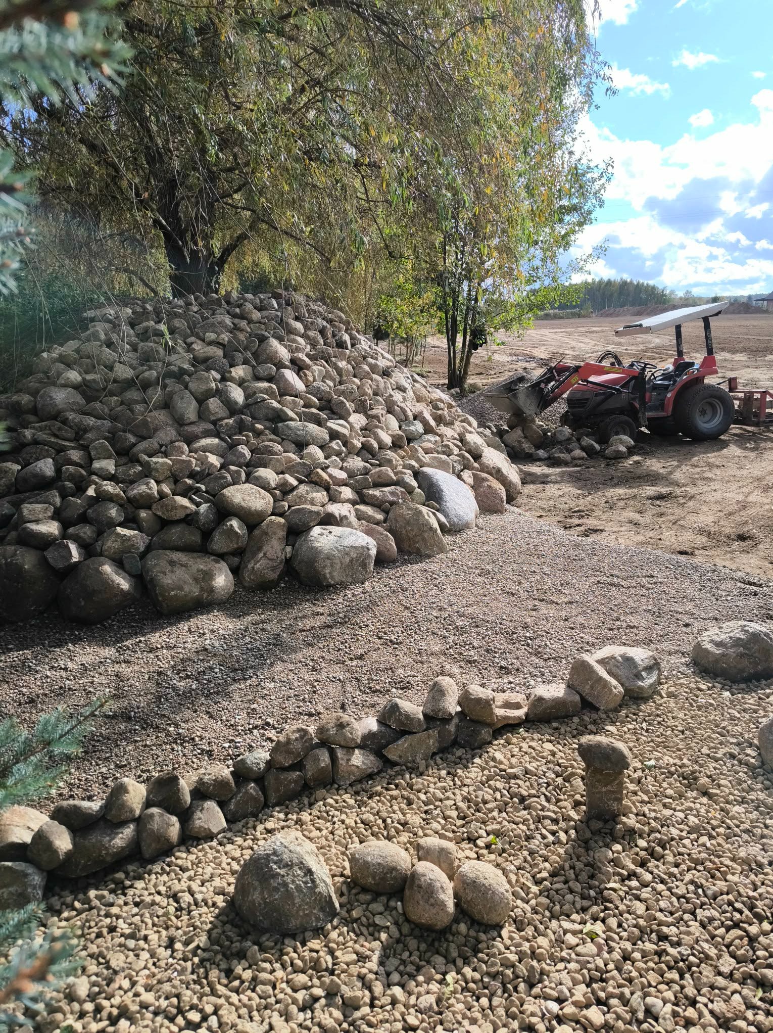 Układanie kamieni ozdobnych w ogrodzie z użyciem traktora. Widoczny kopiec z kamieni, ścieżka z drobnego żwiru i obrzeże z kamieni. Słoneczny dzień.