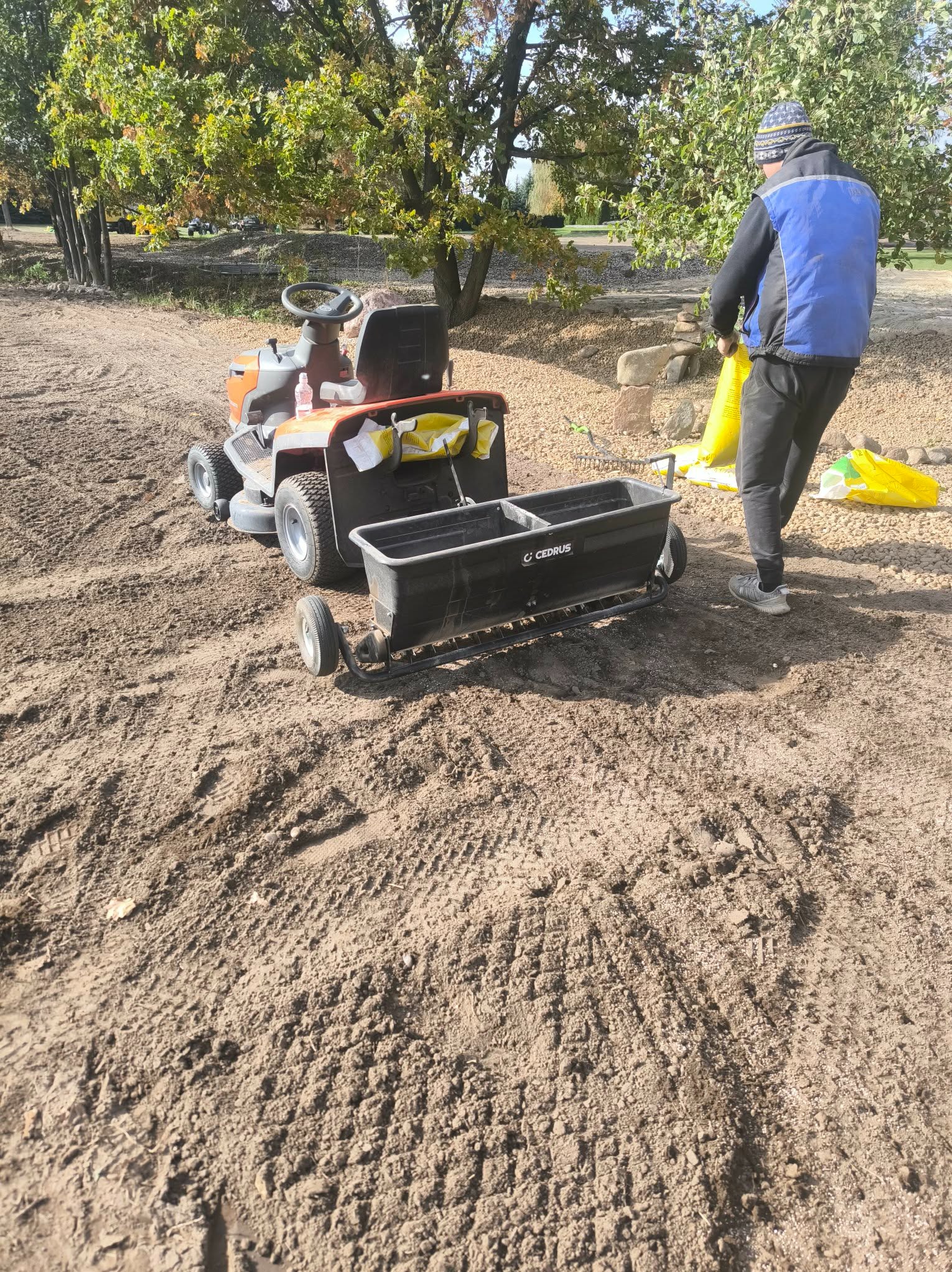 Traktorek ogrodowy z siewnikiem i pracownik rozsypujący nawóz na przygotowanym podłożu. Widoczne ślady opon na ziemi i drzewa w tle.