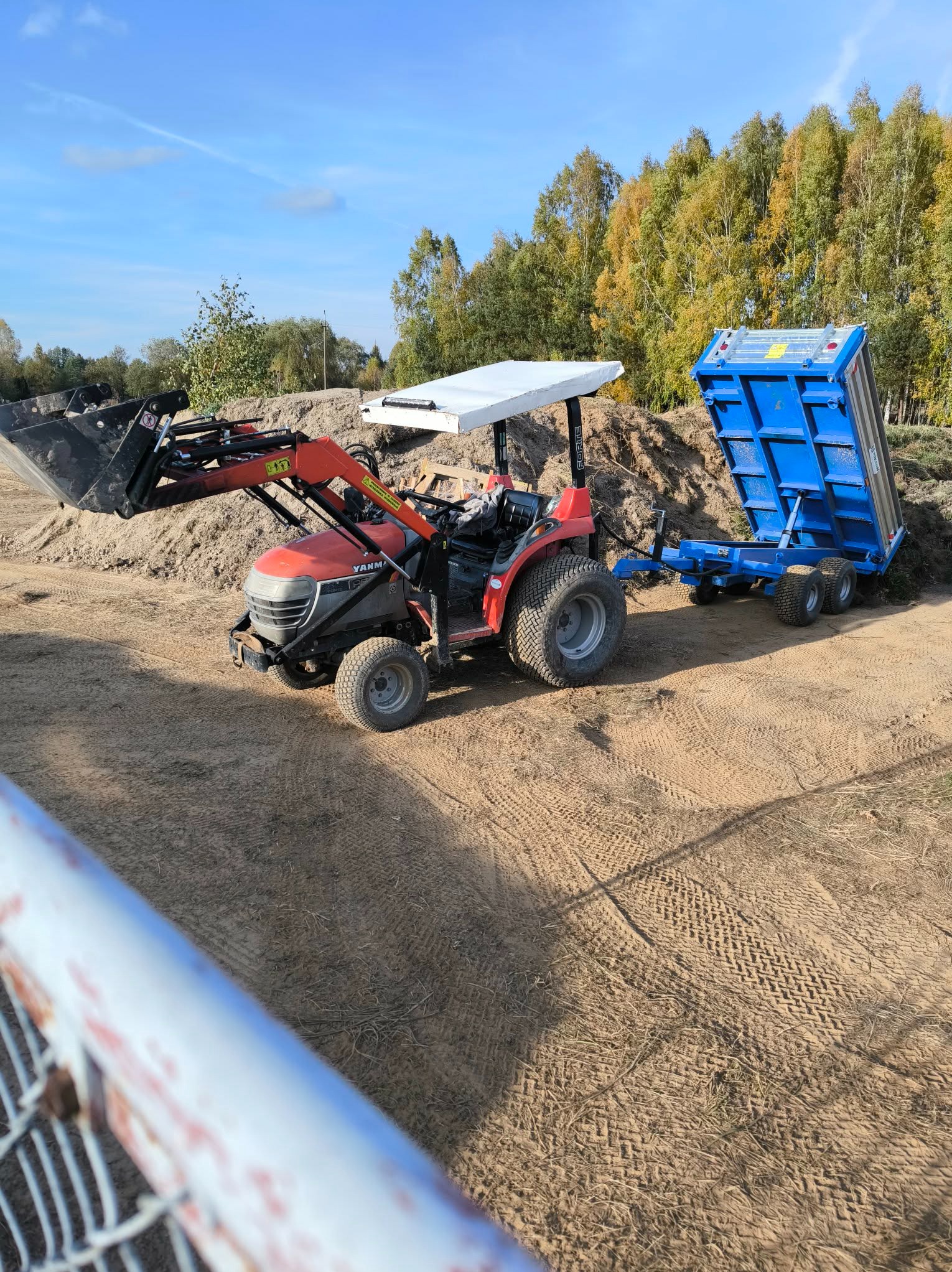 Czerwony traktor Yanmar z przyczepą wywrotką na piaszczystym terenie, w tle drzewa i błękitne niebo. Widoczne ślady opon na ziemi.