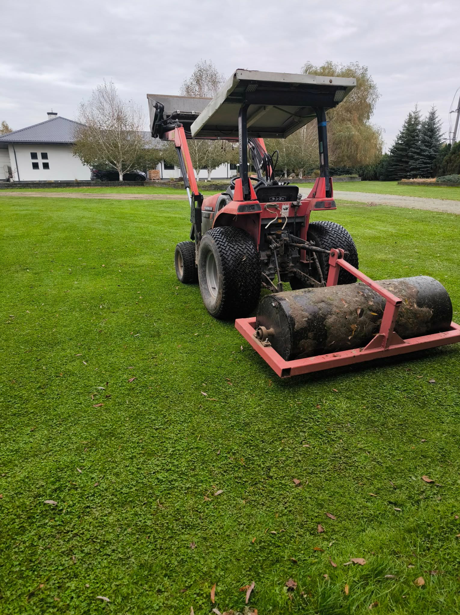 Czerwony traktor z wałem ugniatającym na zielonym trawniku, w tle dom z szarą dachówką i drzewa. Widok z tyłu na maszynę do pielęgnacji trawnika.