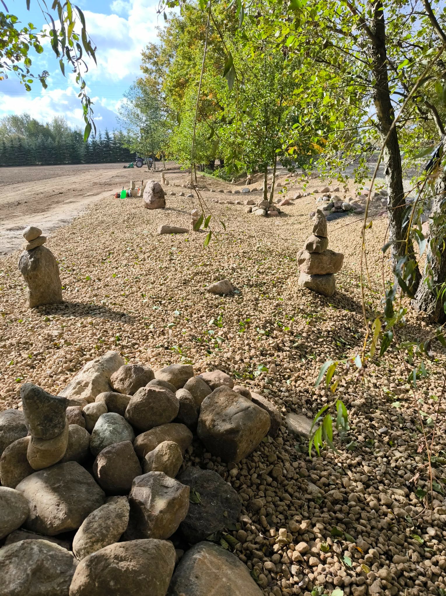 Ogród z kamiennym żwirem, ułożone piramidki z kamieni, drzewa w tle, traktor i narzędzia ogrodnicze. Dekoracyjny element ogrodu.