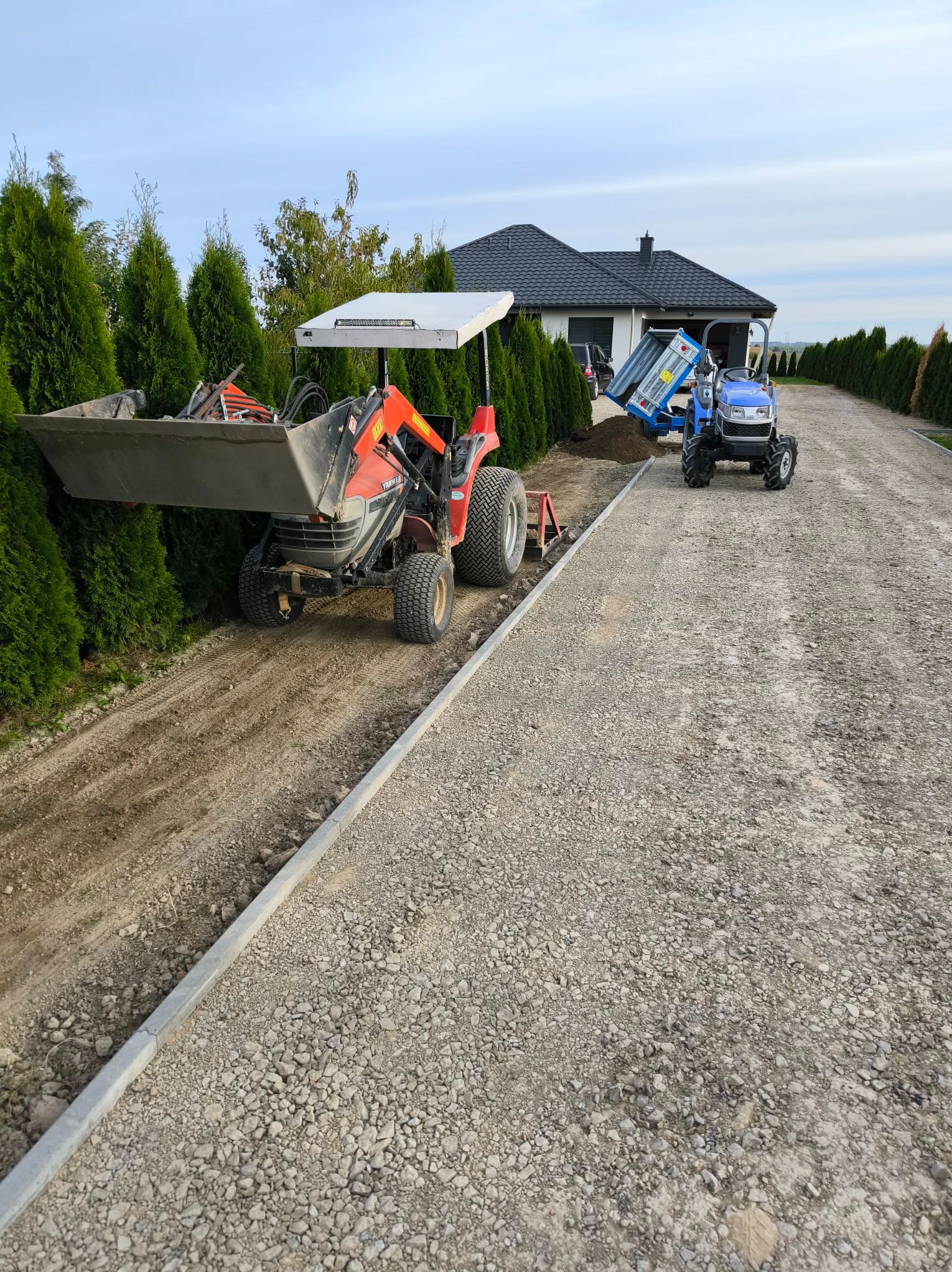 Dwa traktory na żwirowej drodze w trakcie prac ziemnych. Jeden z łyżką, drugi z przyczepą wysypuje ziemię. W tle dom i tuje.