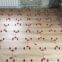 Dariusz Pękalski 5 7 4 3 5 7 8 9 1 - Podłoga w trakcie układania płytek imitujących drewno, z użyciem czerwonych klipsów poziomujących, widoczne surowe ściany i fragment zabudowy kuchennej.