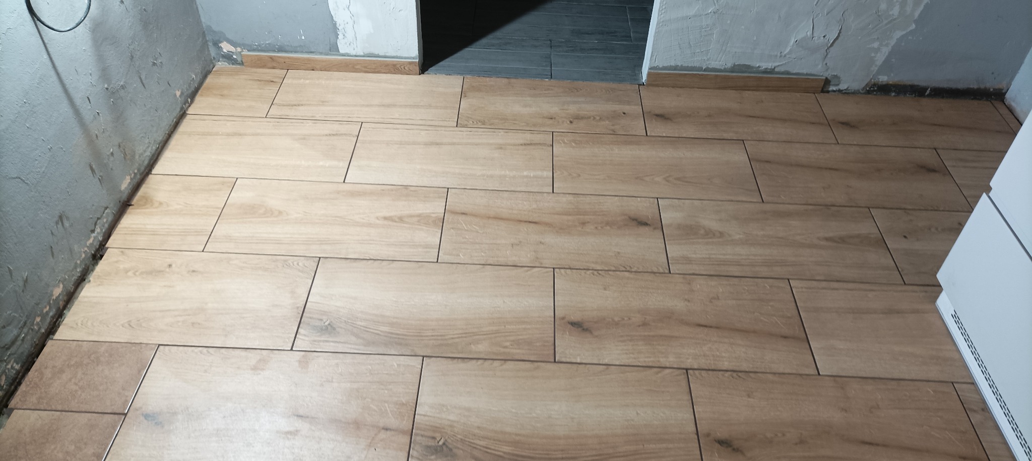 Ułożone podłużne płytki podłogowe imitujące drewno w trakcie wykańczania łazienki, widoczne surowe ściany i fragment progu z szarą płytką.