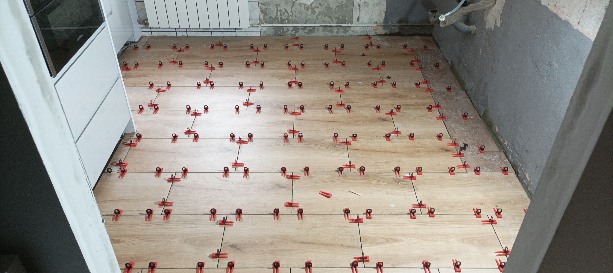 Podłoga w trakcie układania płytek imitujących drewno, z użyciem czerwonych klipsów poziomujących, widoczne surowe ściany i fragment zabudowy kuchennej.