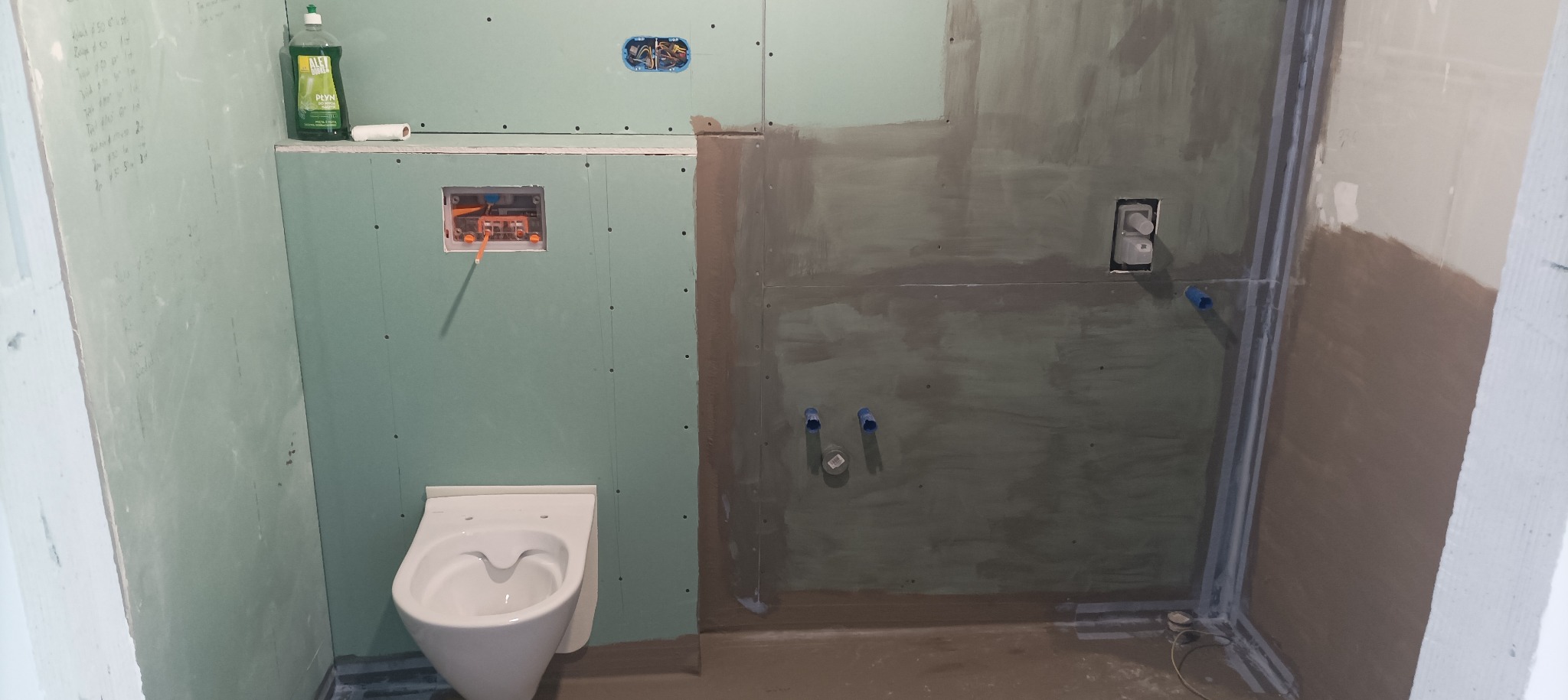 Łazienka w trakcie prac wykończeniowych: zamontowana toaleta wisząca, ściany z płyt gipsowo-kartonowych z widocznymi instalacjami, częściowo zagruntowane, butelka płynu do naczyń na półce.