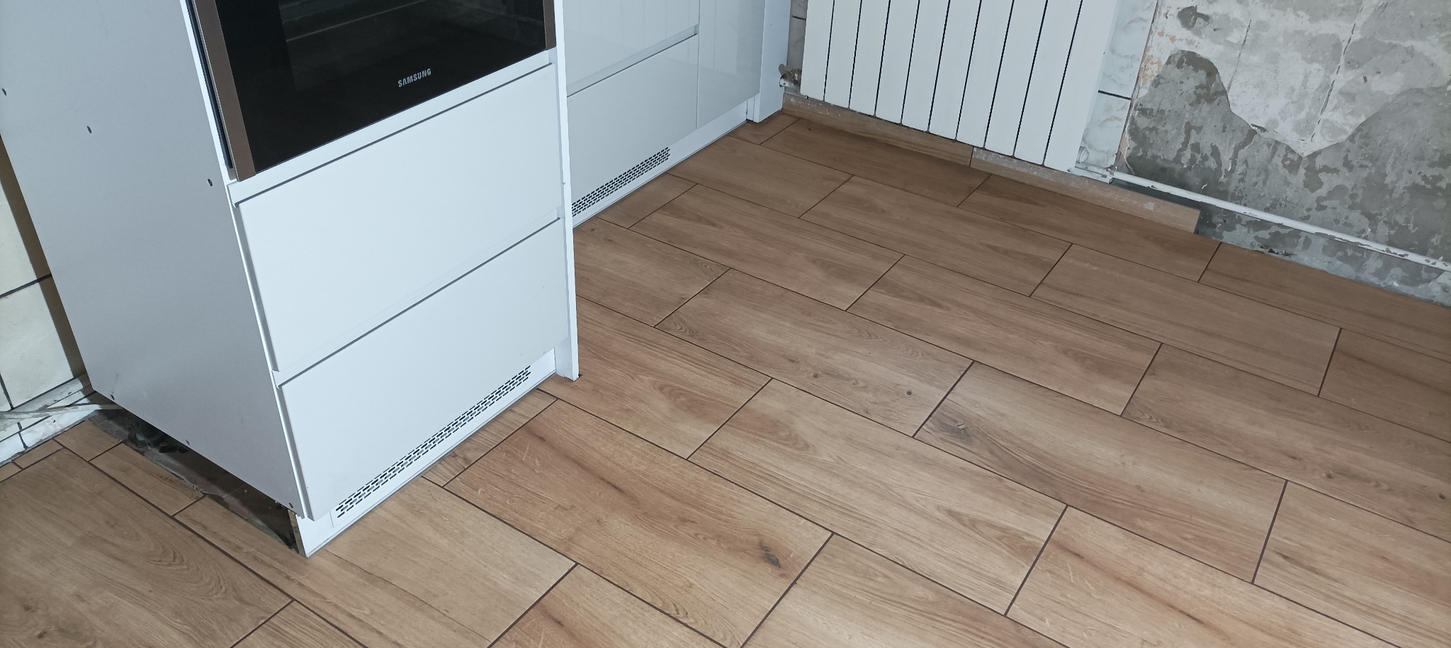 Ułożona podłoga z płytek imitujących drewno w kuchni, widoczny fragment białej zabudowy kuchennej i grzejnik na tle surowej ściany w trakcie remontu.
