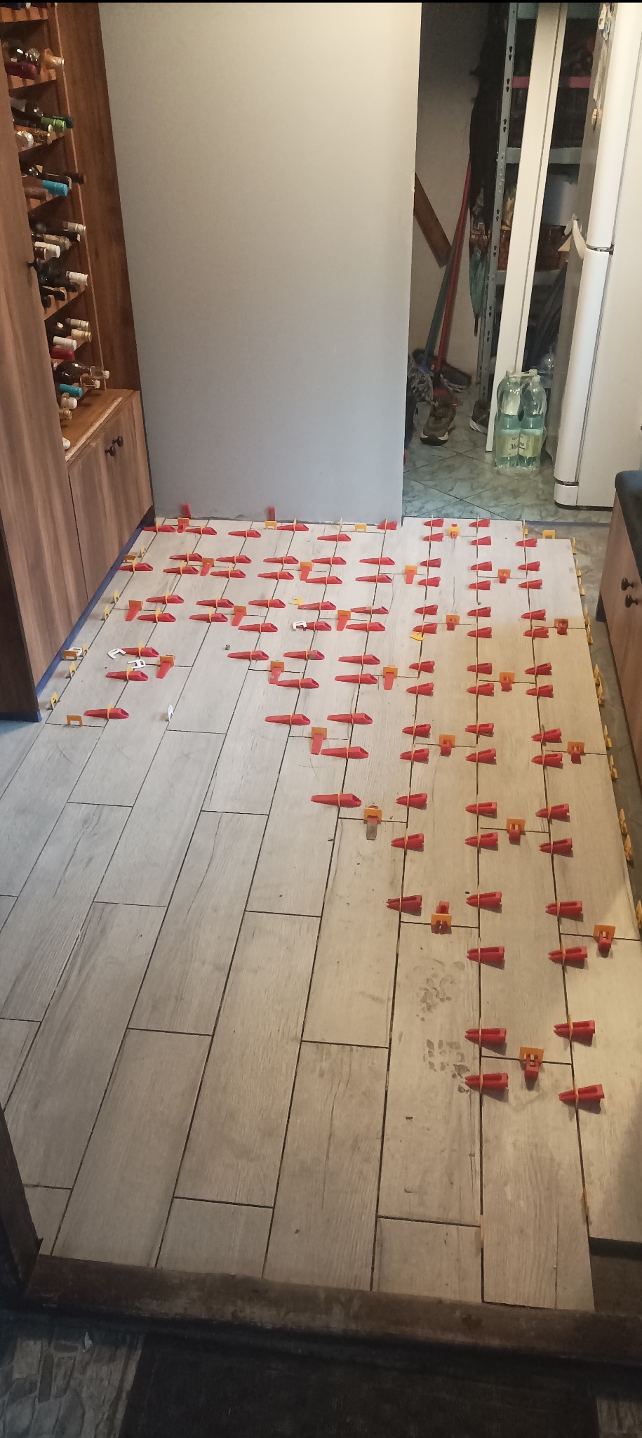 Układanie płytek podłogowych imitujących drewno, z użyciem czerwonych i żółtych klipsów poziomujących, widok na fragment pomieszczenia z szafką na wino i otwartym przejściem do zagraconego...