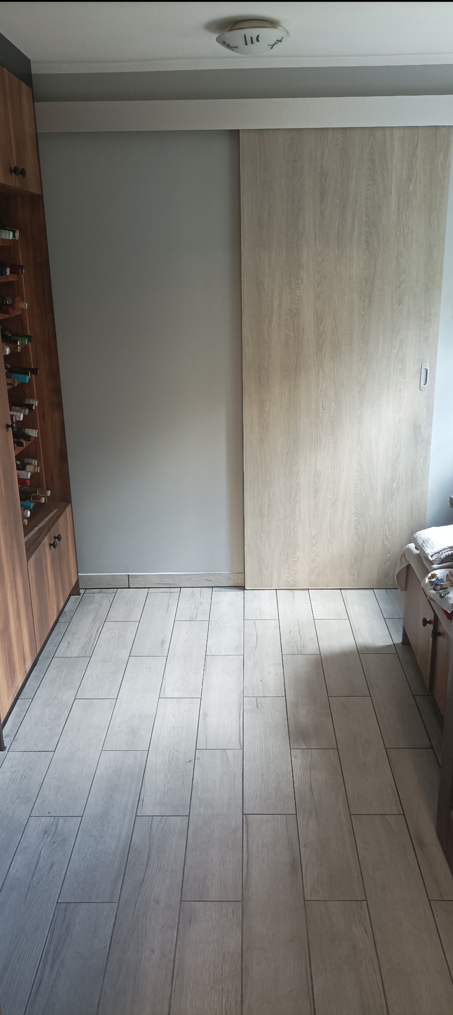 Jasnoszare, podłużne płytki podłogowe imitujące drewno, ułożone w łazience z przesuwnymi drzwiami i drewnianą szafką z półkami na wino.