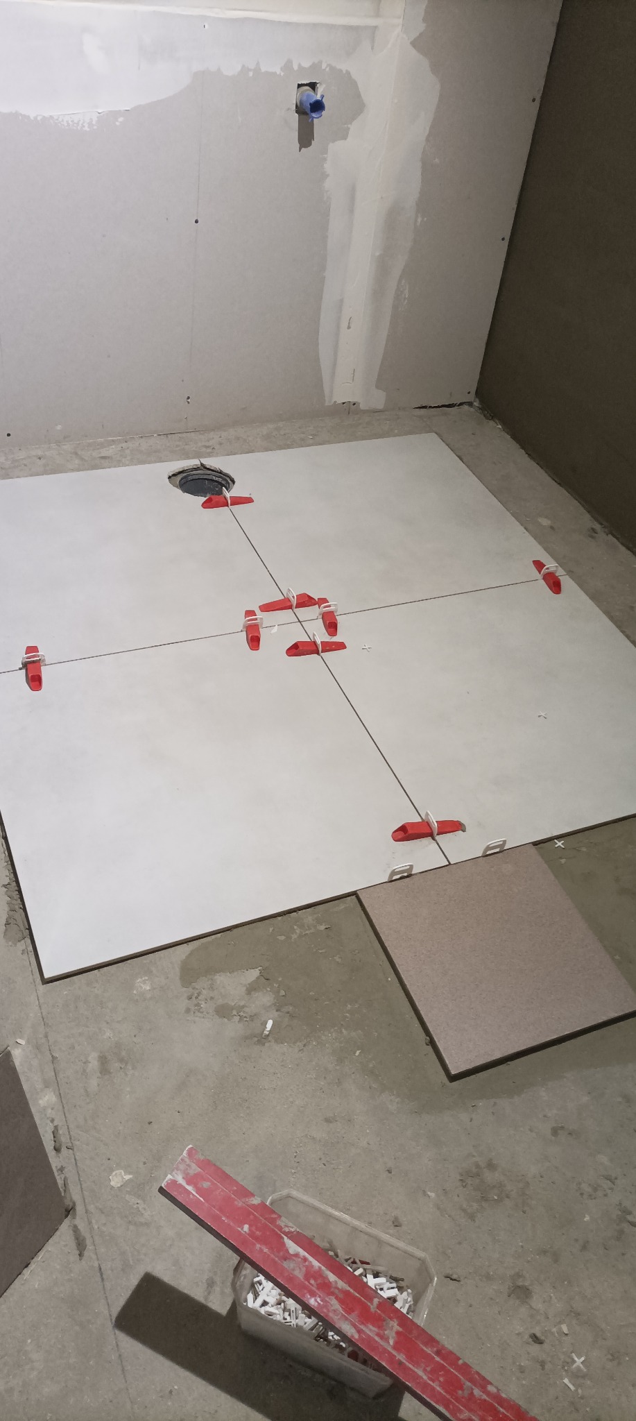 Układanie jasnych płytek podłogowych w łazience z użyciem systemu poziomowania płytek (czerwone klipsy i kliny), widoczny odpływ podłogowy, częściowo wykończone ściany.