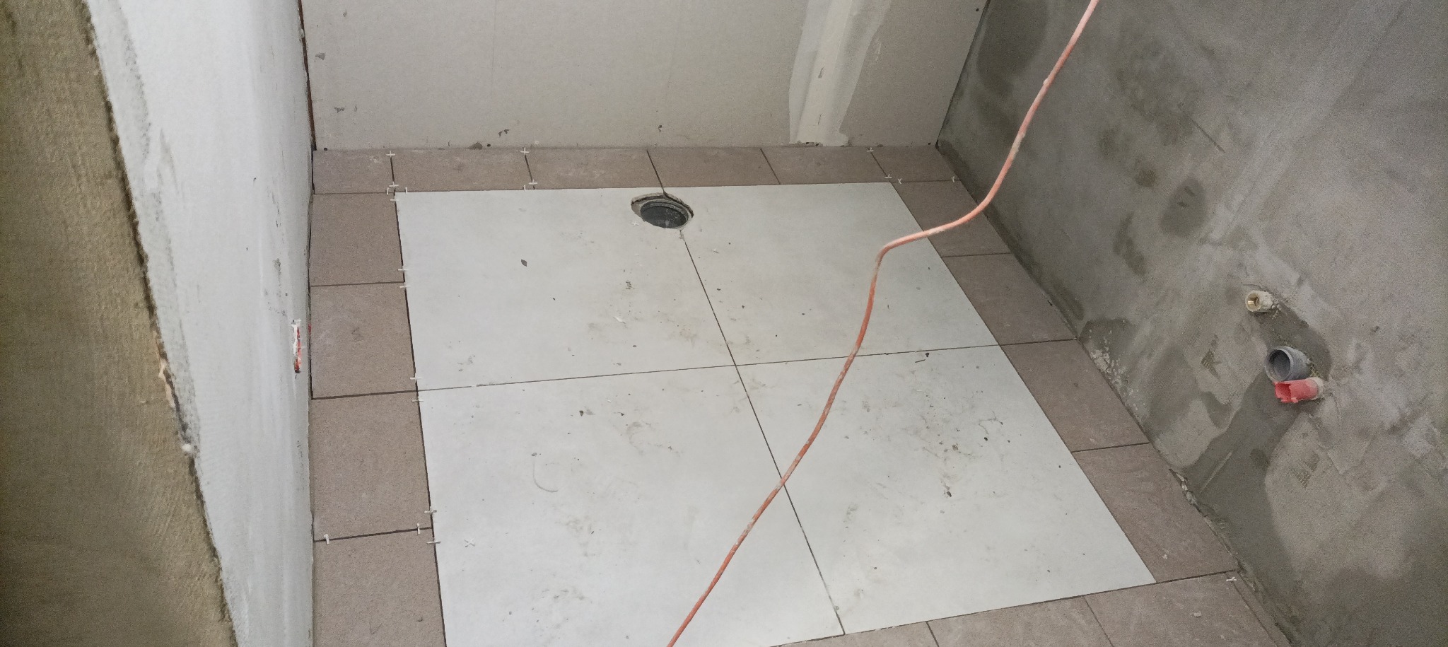 Fragment łazienki w trakcie układania płytek podłogowych: białe płytki ułożone na środku pomieszczenia, brązowe płytki przy ścianach, widoczne krzyżyki dystansowe, odpływ liniowy w podłodze, surowe...