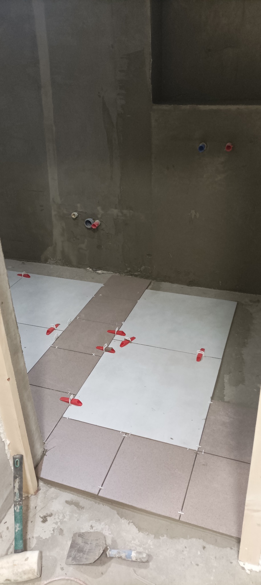 Układanie jasnych i beżowych płytek podłogowych w łazience z użyciem czerwonych klinów poziomujących, widoczne przyłącza wodne na surowej ścianie.