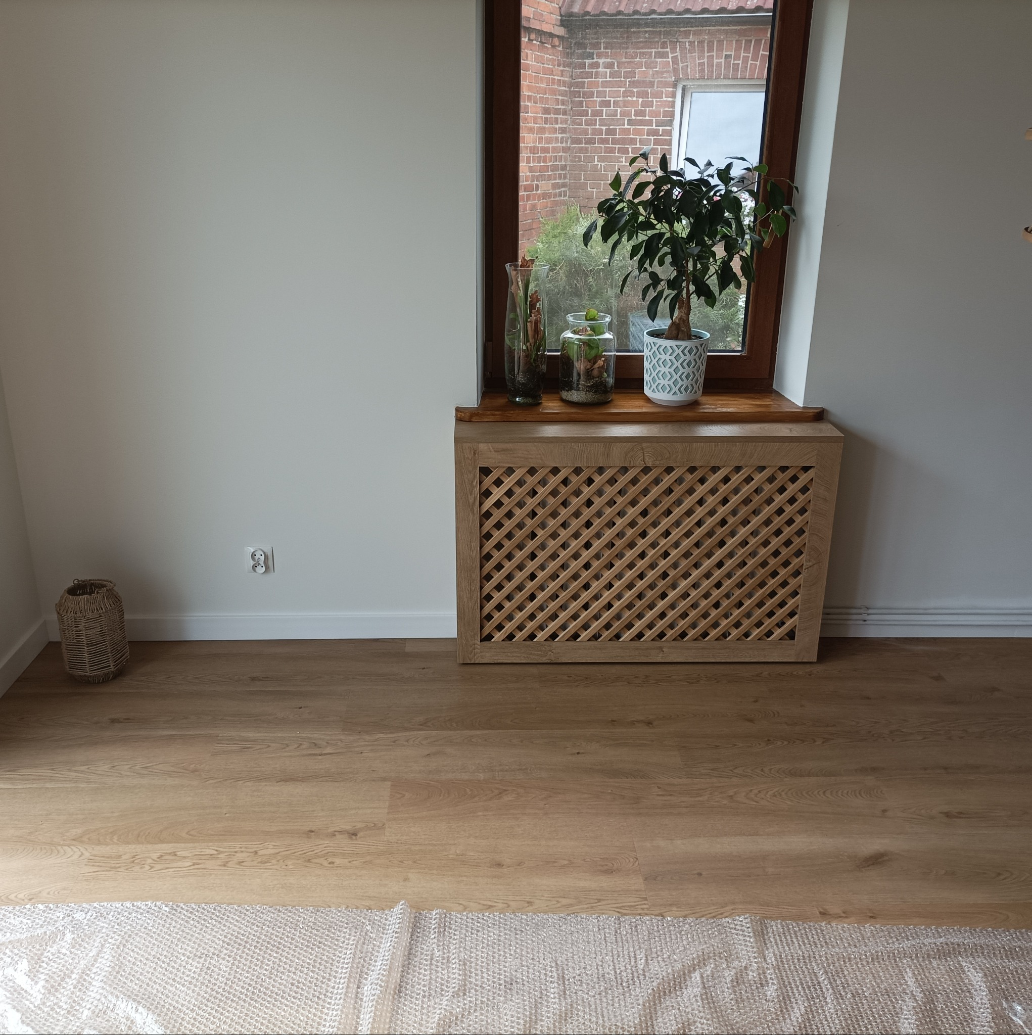 Drewniana osłona grzejnika z kratką w jasnym wnętrzu, na parapecie rośliny w szklanych naczyniach i doniczce, podłoga wyłożona panelami, częściowo przykryta folią bąbelkową.