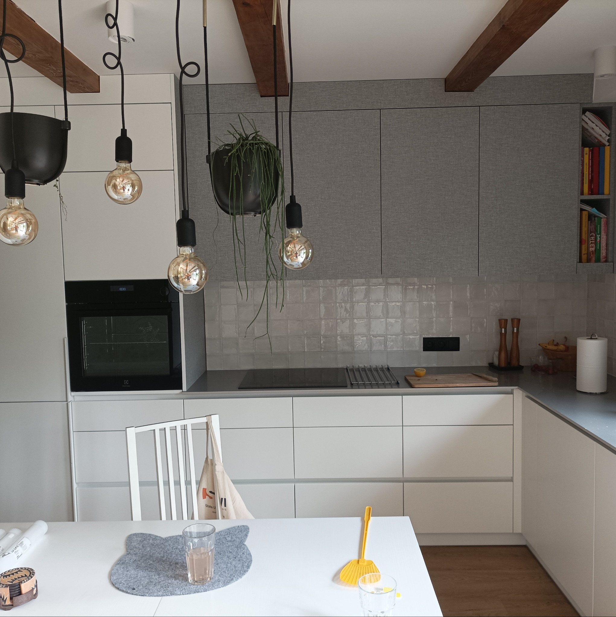 Nowoczesna kuchnia z szarymi szafkami, białymi frontami, drewnianymi belkami na suficie i wiszącymi lampami z żarówkami Edisona oraz roślinami w czarnych donicach. Na blacie leży cytryna. Na stole...