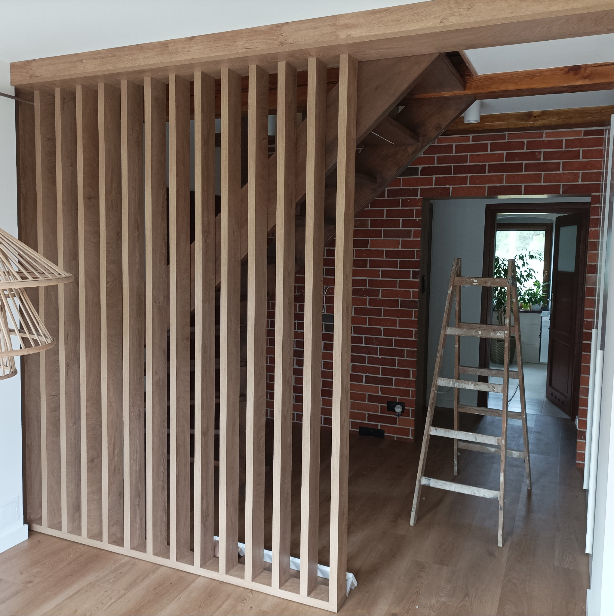 Drewniana ścianka działowa z pionowych listew, widoczna na tle ceglanej ściany i drewnianych schodów, z drabina opartą o ścianę.