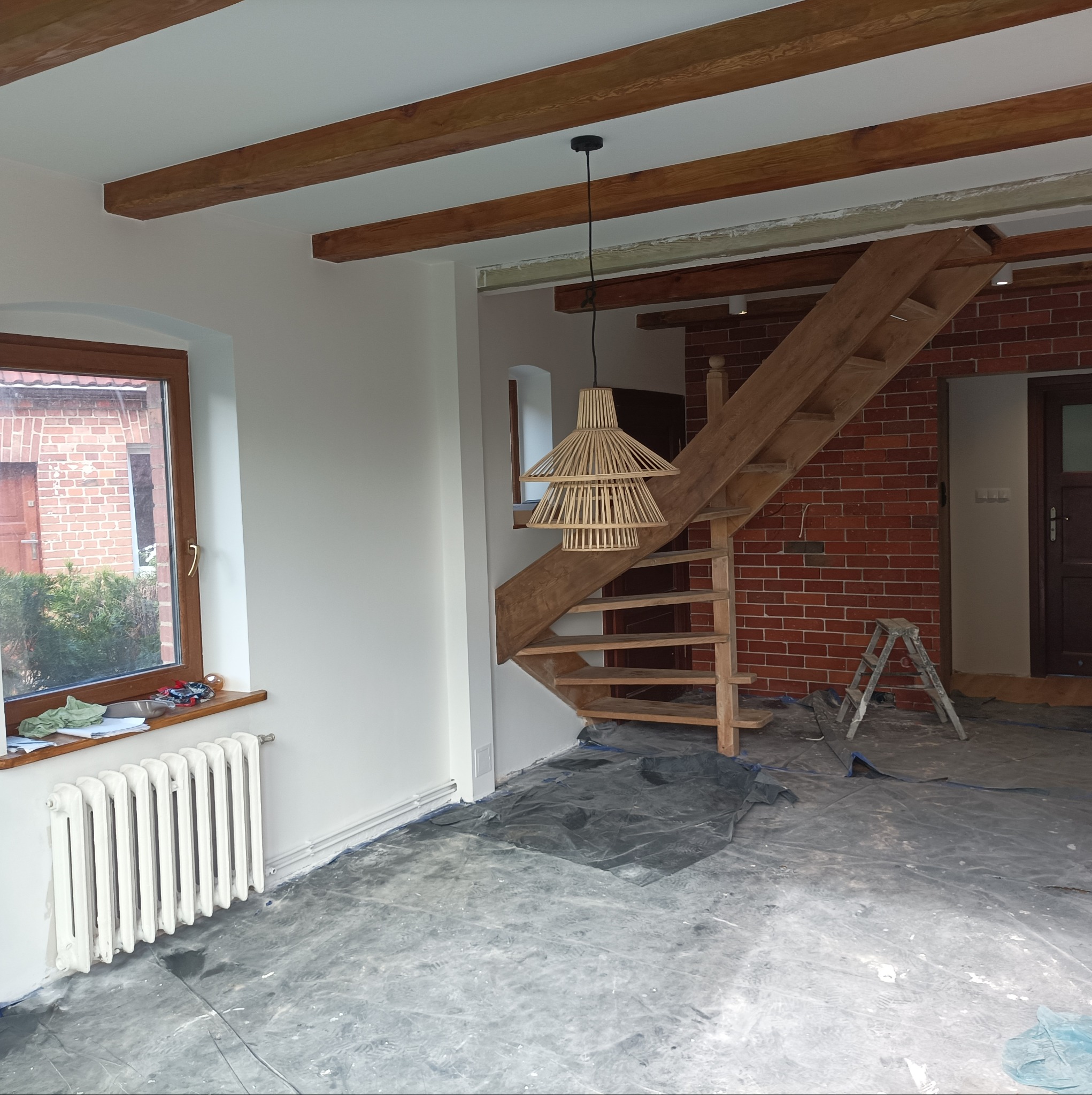 Wnętrze w trakcie remontu: surowe drewniane schody, lampa z naturalnych materiałów, odsłonięta cegła na ścianie, zabezpieczona folią podłoga, grzejnik pod oknem.