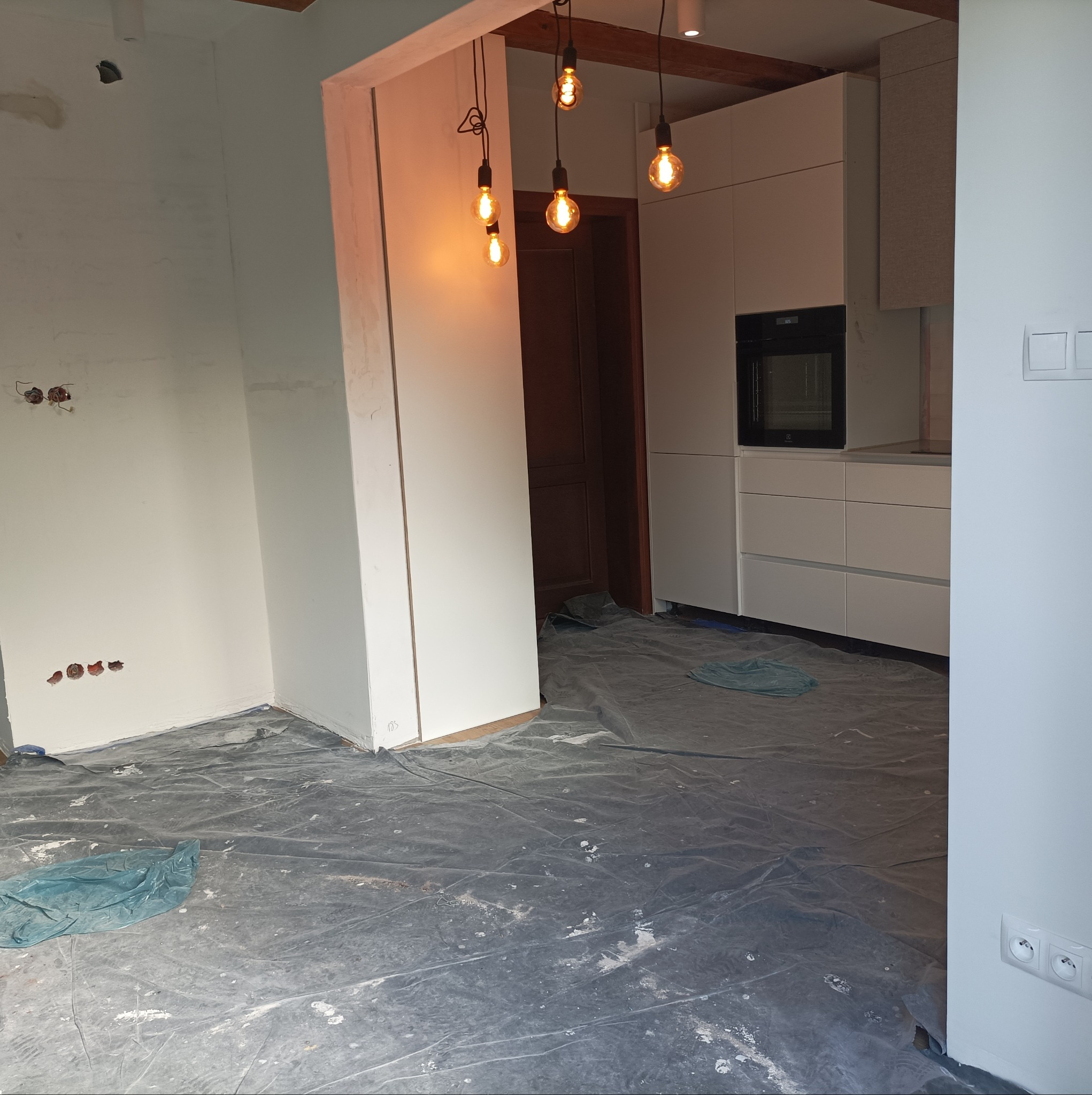 Remont kuchni w trakcie: białe ściany po gładzi, odkryte gniazdka elektryczne, podłoga zabezpieczona folią malarską, w tle zabudowa kuchenna z piekarnikiem i wiszące żarówki.