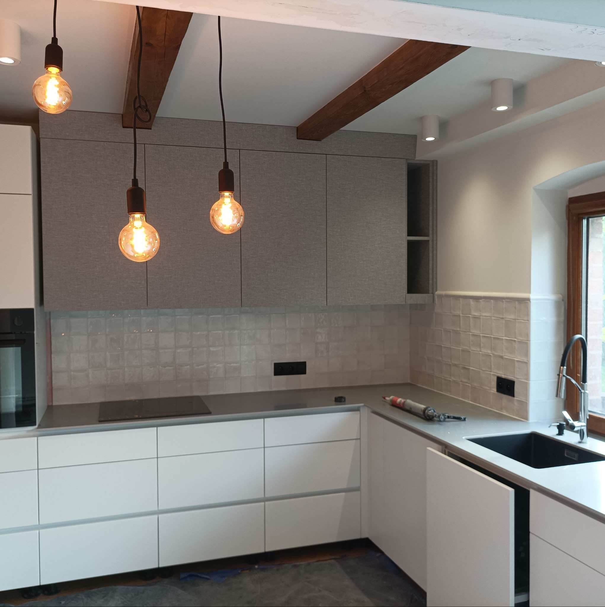 Nowoczesna kuchnia z szarymi szafkami górnymi o teksturowanym wykończeniu, białymi szafkami dolnymi, kwadratowymi płytkami ściennymi, drewnianymi belkami sufitowymi i trzema wiszącymi lampami...