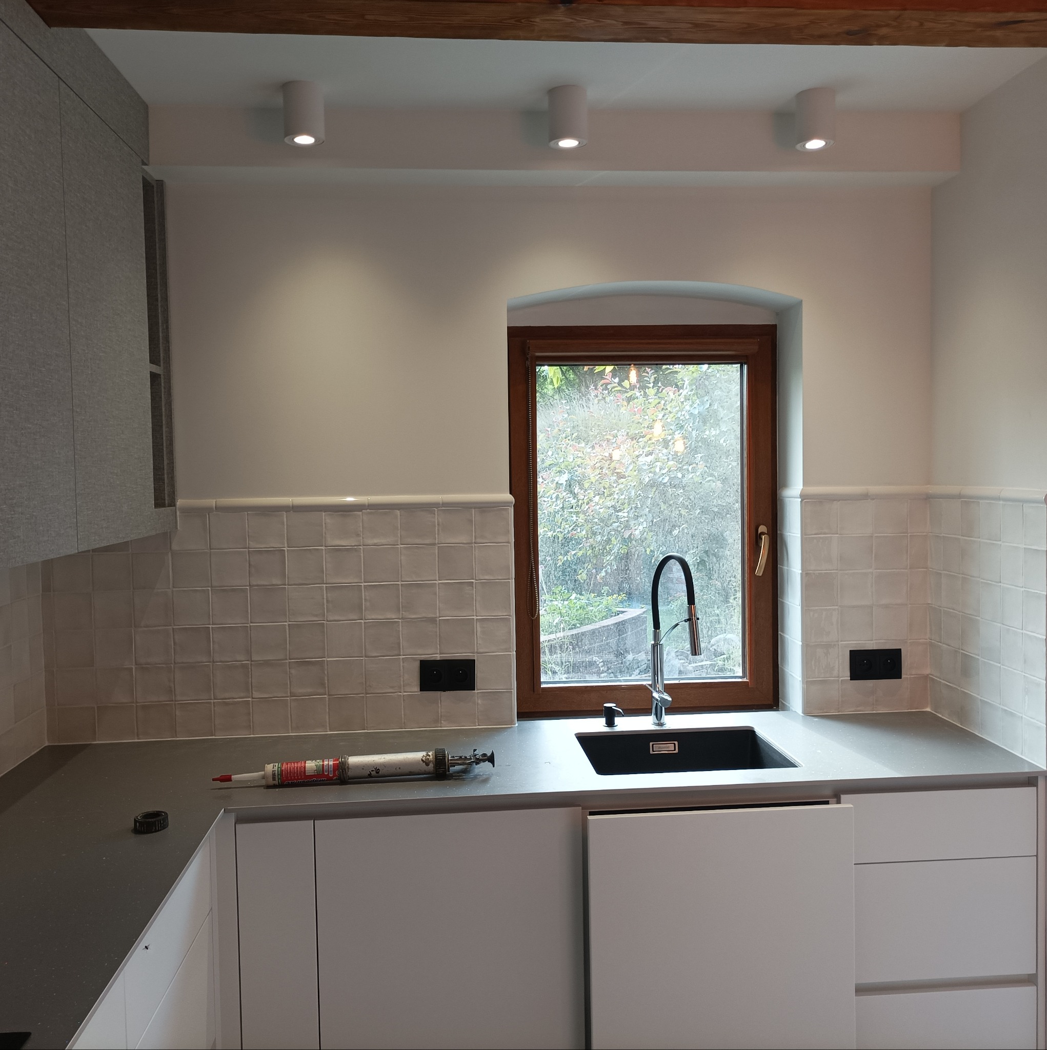 Wykończona kuchnia z białymi płytkami kwadratowymi, drewnianym oknem, czarnym zlewem i tubą silikonu na blacie, widok na zieleń za oknem.