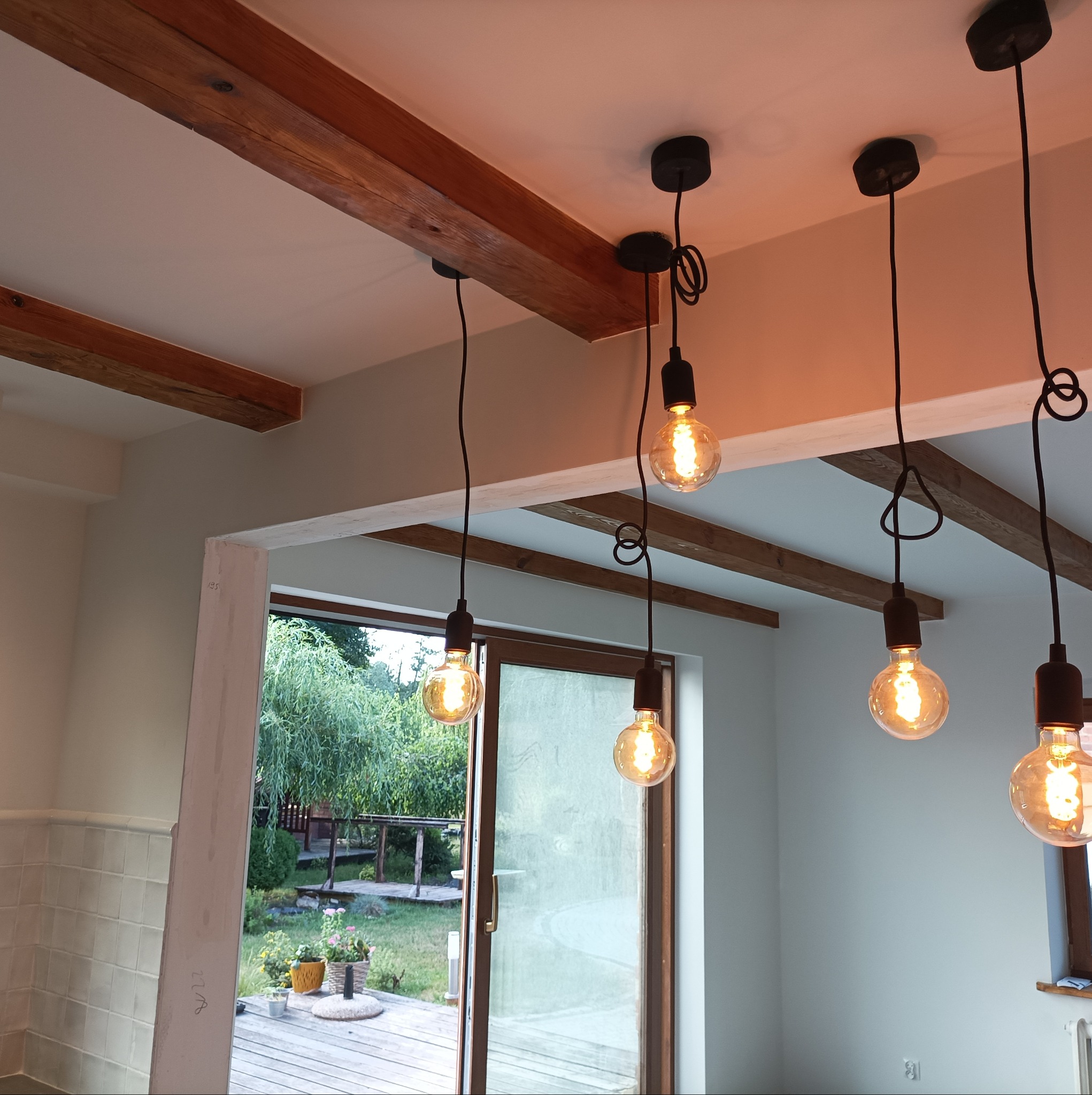 Wnętrze pokoju z drewnianymi belkami na suficie i czterema wiszącymi lampami z żarówkami Edisona, widok na ogród przez przesuwne drzwi balkonowe.