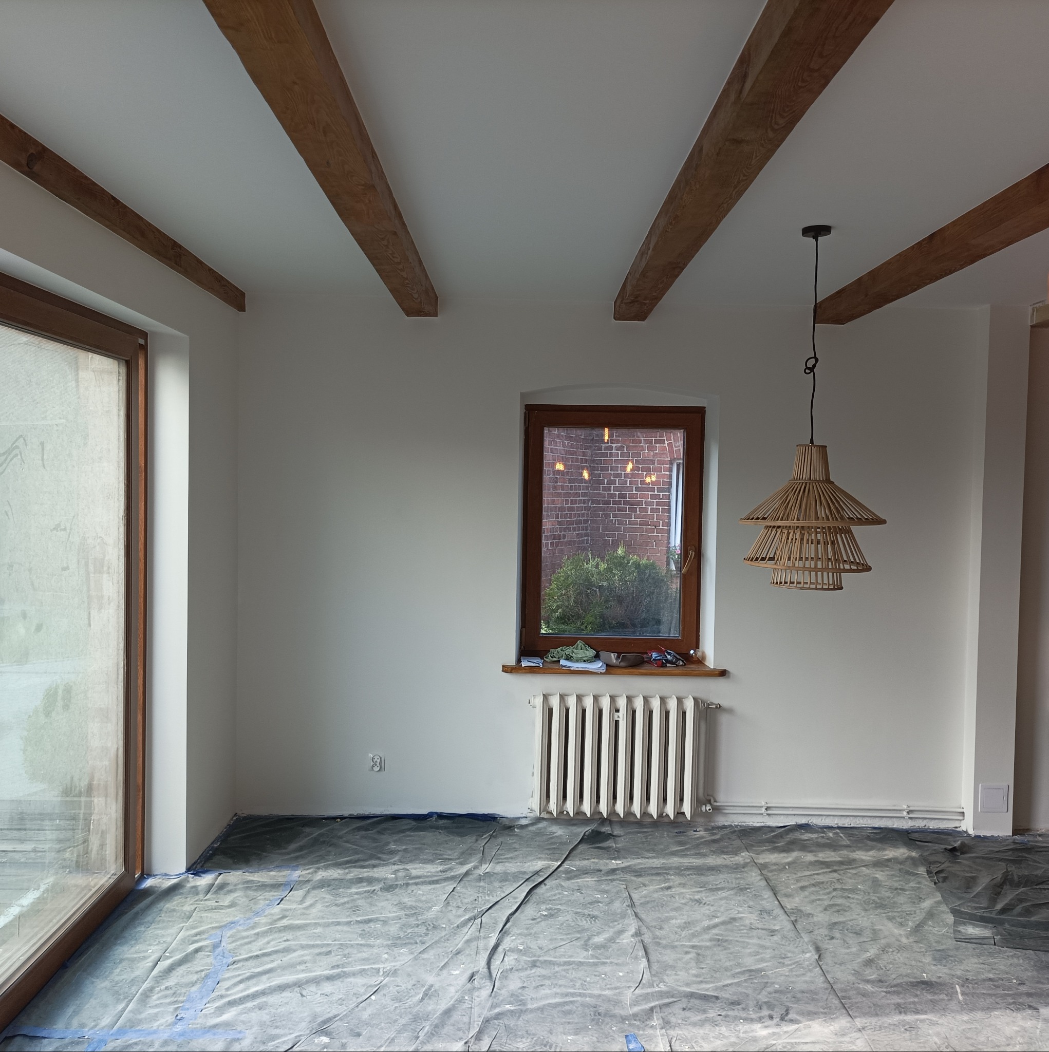 Pokój w trakcie remontu: zabezpieczona folią podłoga, świeżo pomalowane ściany na biało, drewniane belki stropowe, okno z widokiem na ceglany budynek, wisząca lampa z naturalnego materiału.