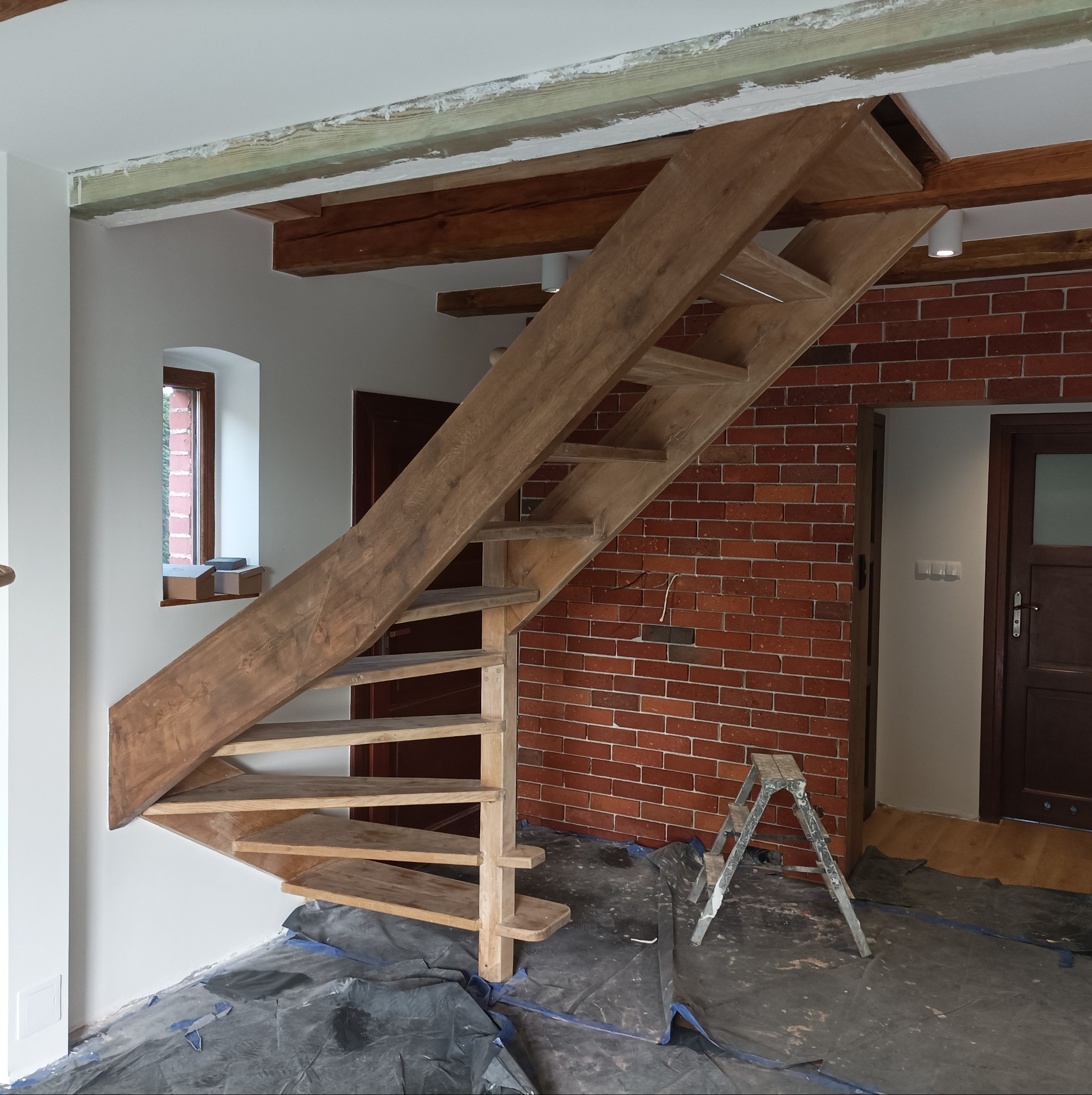 Drewniane schody spiralne w trakcie montażu, widoczna drabina malarska, zabezpieczona folią podłoga, ceglana ściana w tle.