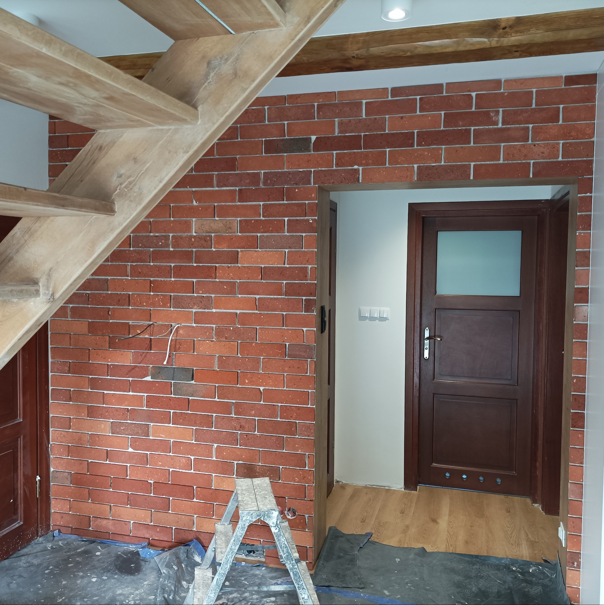 Montaż cegieł dekoracyjnych na ścianie w korytarzu, widoczne drewniane schody, drzwi z szybą i rozłożona folia ochronna na podłodze.