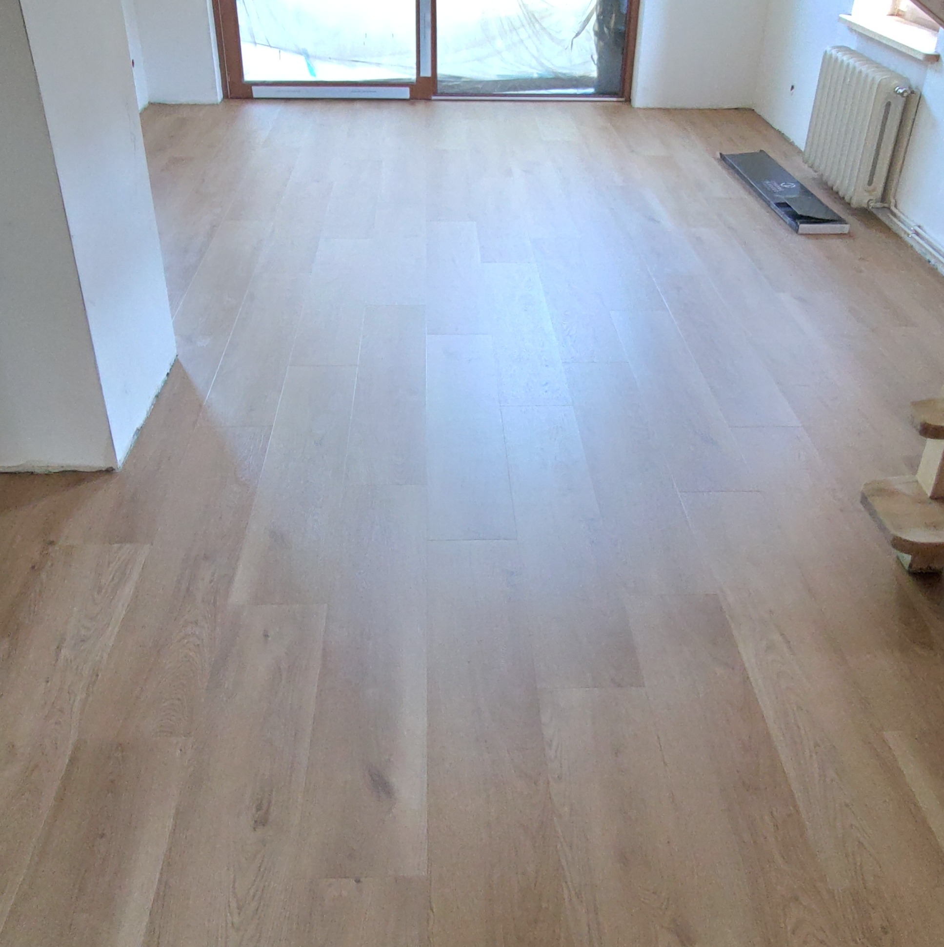 Jasne panele podłogowe ułożone w pokoju, widoczny fragment grzejnika i okna zabezpieczonego folią malarską, oraz karton po panelach.