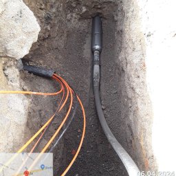 FPUH ATOM Adrian Tomczyk - Wąski wykop w ziemi z widocznym wiertłem kierunkowym i pomarańczowymi kablami, widoczny fragment mapy z lokalizacją.