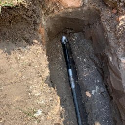 FPUH ATOM Adrian Tomczyk - Wąski wykop w ziemi z odsłoniętym murem ceglanym, wewnątrz rura w osłonie z głowicą wiercącą na końcu.