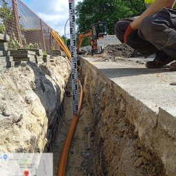 FPUH ATOM Adrian Tomczyk - Pomarańczowe rury ułożone w wykopie, pracownik mierzy głębokość za pomocą łaty niwelacyjnej, w tle mini koparka i ogrodzenie z siatki.