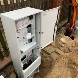 FPUH ATOM Adrian Tomczyk - Biała szafka telekomunikacyjna z otwartymi drzwiami, umieszczona w wykopie ziemnym, widoczna koparka w tle, obok drewnianego płotu i ceglanego słupka.