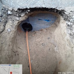 FPUH ATOM Adrian Tomczyk - Wykop w ziemi z widoczną rurą osłonową i pomarańczowym przewodem, zabezpieczony niebieską folią, położony w pobliżu betonowej nawierzchni.