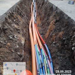 FPUH ATOM Adrian Tomczyk - Wykop w ziemi z ułożonymi rurami światłowodowymi, pomiar głębokości za pomocą poziomicy i łaty niwelacyjnej, widoczny pomarańczowy kask ochronny i dane geolokalizacyjne.