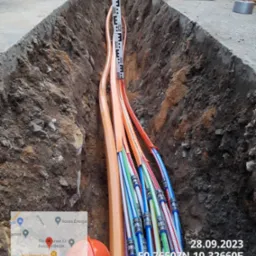 Wykop w ziemi z ułożonymi rurami światłowodowymi, pomiar głębokości za pomocą poziomicy i łaty niwelacyjnej, widoczny pomarańczowy kask ochronny i dane geolokalizacyjne.