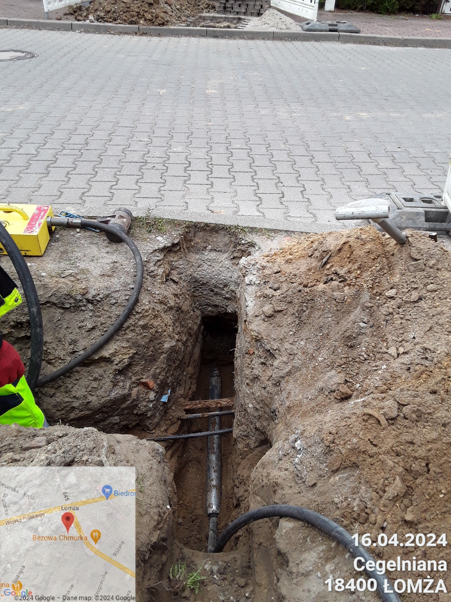 Wykop w ziemi na ulicy, widoczne rury i sprzęt do robót ziemnych, w tle kostka brukowa i data z adresem.
