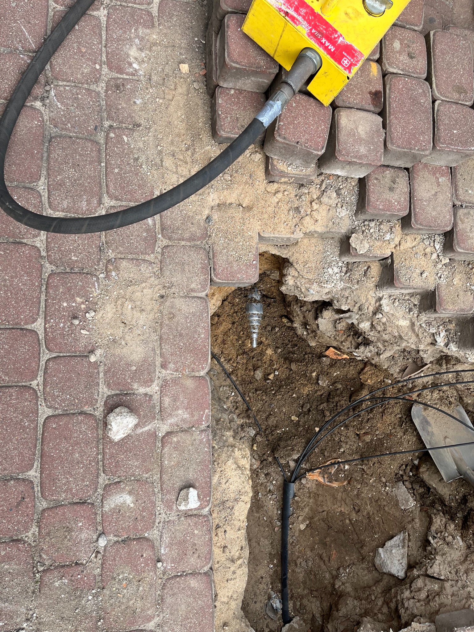 Wiertnica z żółtą głowicą i czarnym przewodem hydraulicznym przy wykopie w kostce brukowej, widoczna instalacja w ziemi i fragment szpadla.