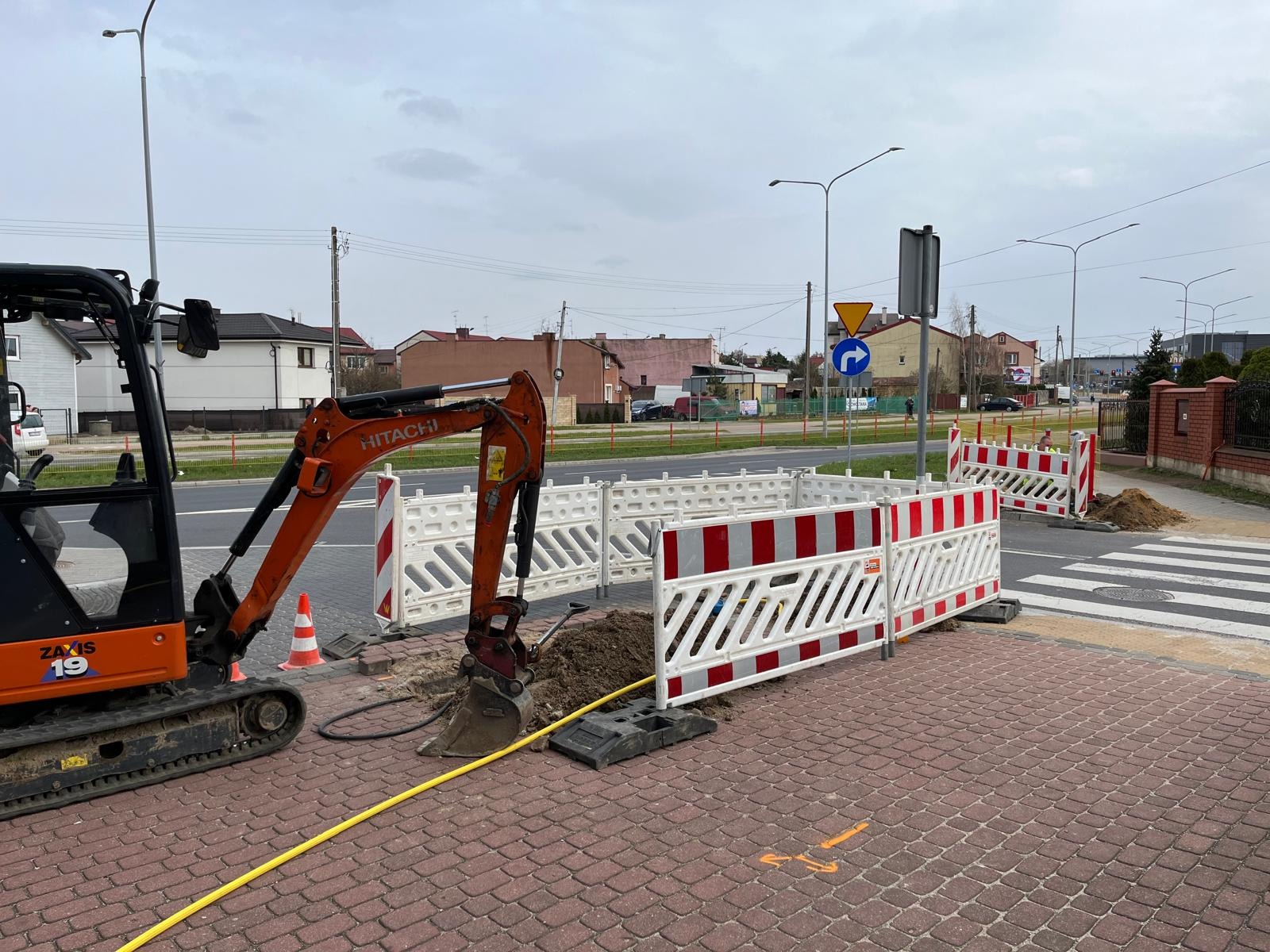 Pomarańczowy minikoparka Hitachi Zaxis 19 przy wykopie zabezpieczonym biało-czerwoną barierką na chodniku z kostki, widoczne oznaczenia kierunku na chodniku, w tle ulica i budynki.