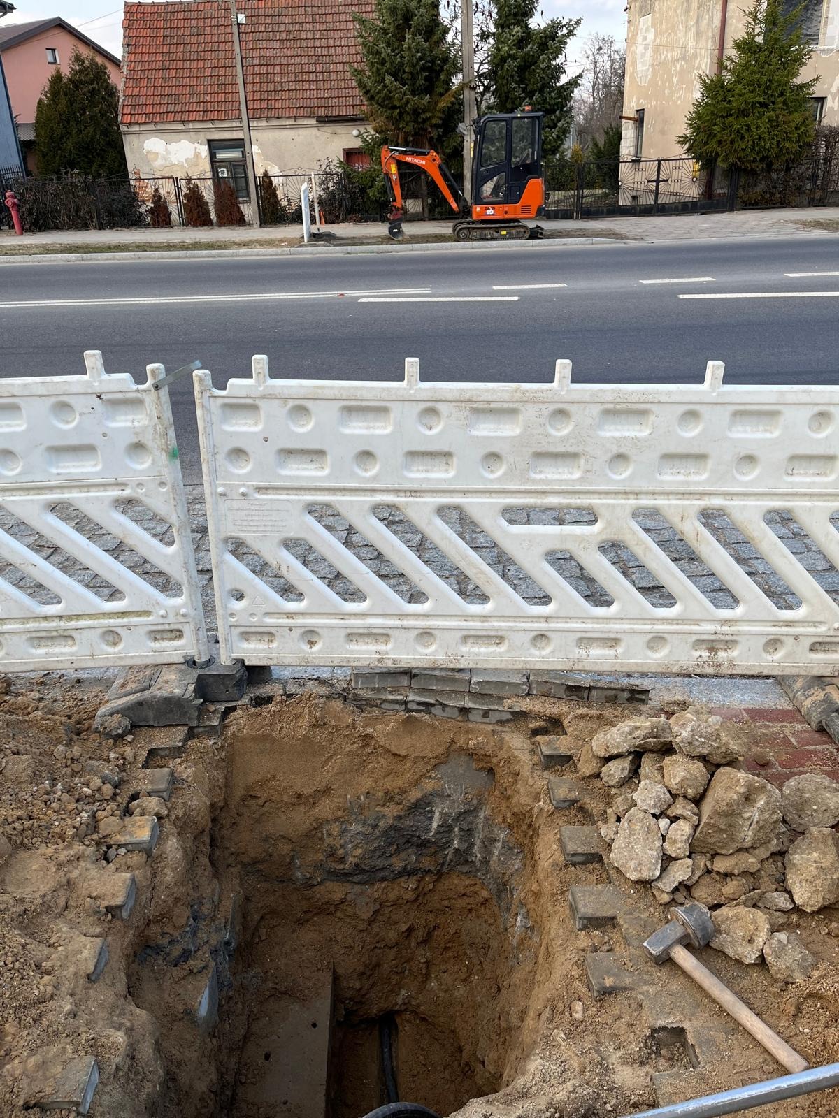 Głęboki wykop w ziemi zabezpieczony białą barierką, z widocznymi przewodami wewnątrz, młotek leżący obok, mała koparka w tle na ulicy.