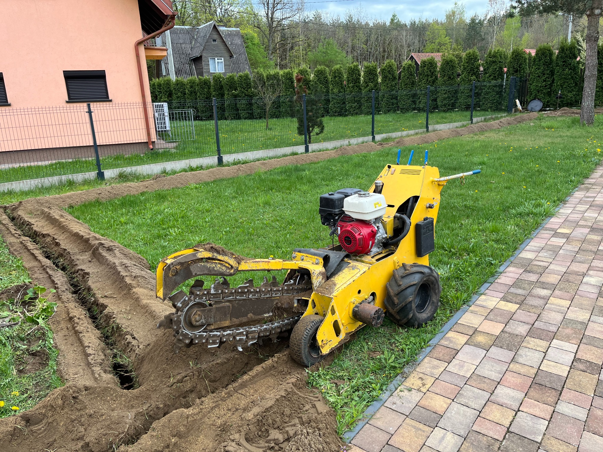 Żółta frezarka łańcuchowa w trakcie wykonywania wąskiego wykopu w trawniku przy posesji z kostką brukową i ogrodzeniem z siatki.