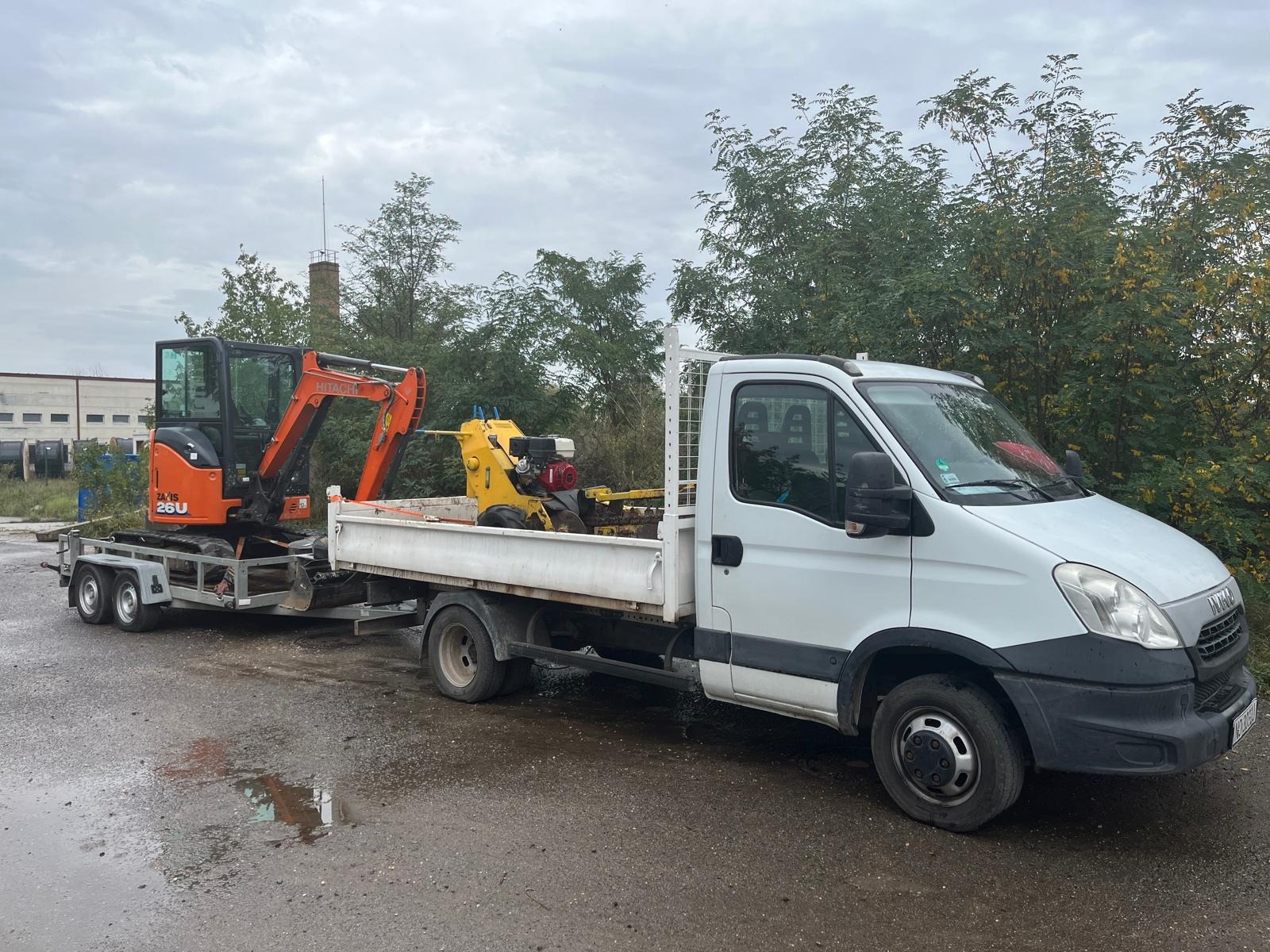 Pomarańczowa mini koparka Hitachi na przyczepie załadowana na białą ciężarówkę Iveco, obok żółta maszyna do robót ziemnych, w tle drzewa i budynek przemysłowy.