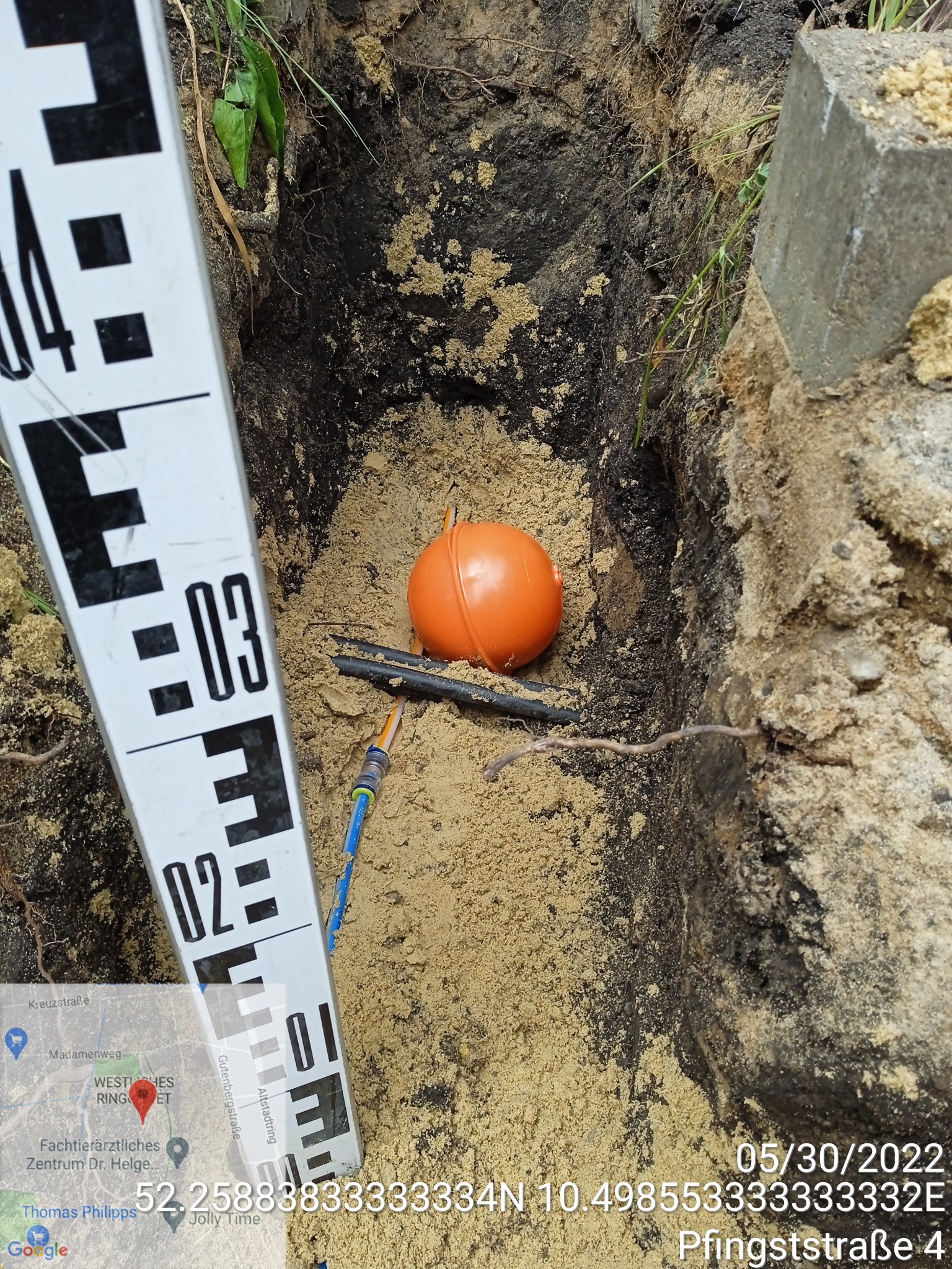 Wykop w ziemi z widoczną pomarańczową kulą, czarnymi kablami i niebieskim przewodem, obok umieszczona miarka geodezyjna, w tle ziemia i betonowy element