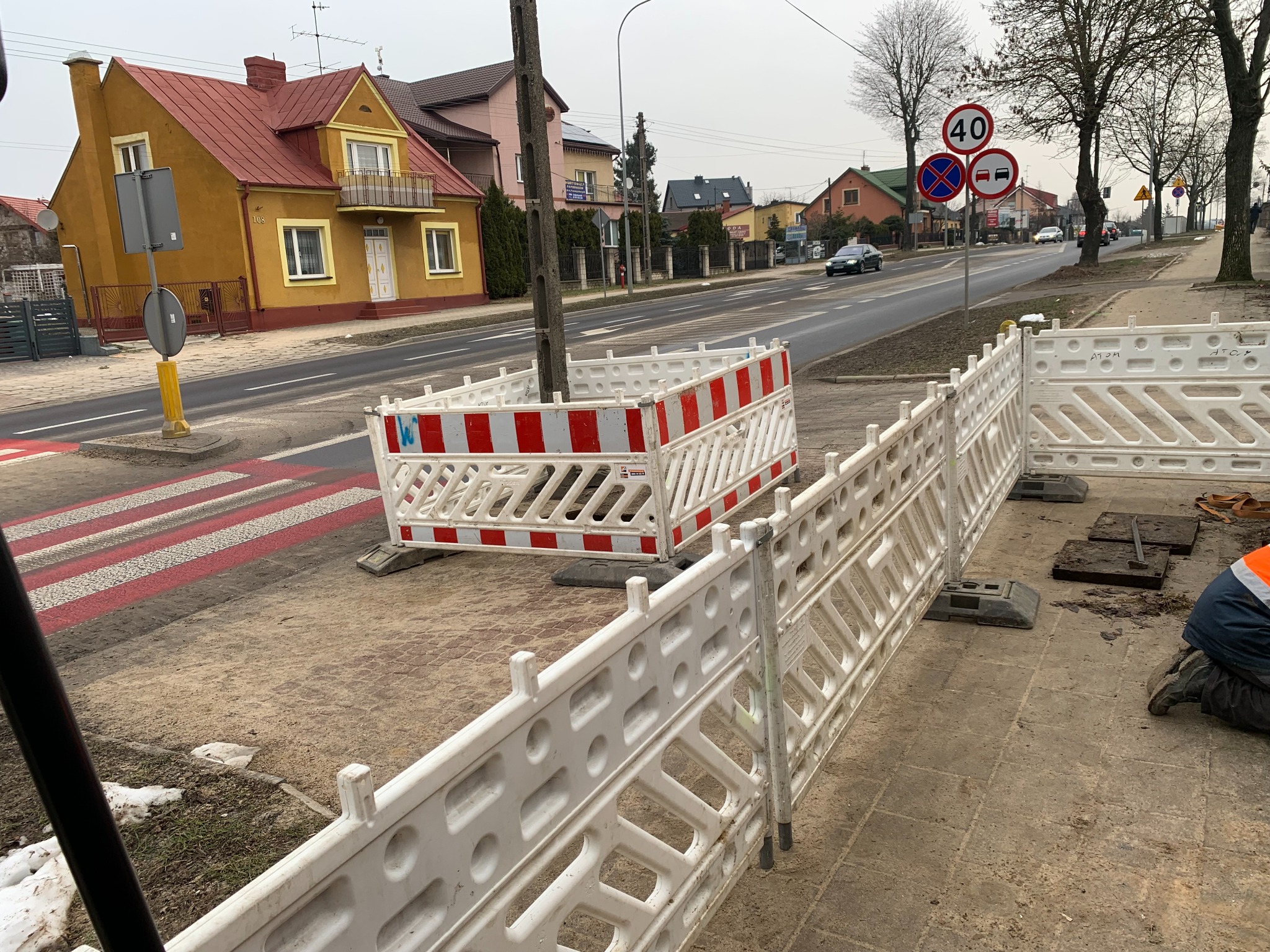 Oznakowanie robót drogowych na chodniku przy przejściu dla pieszych, widoczne barierki ochronne i pracownik w odblaskowej kamizelce przy studzience kanalizacyjnej, w tle zabudowa miejska i znaki...