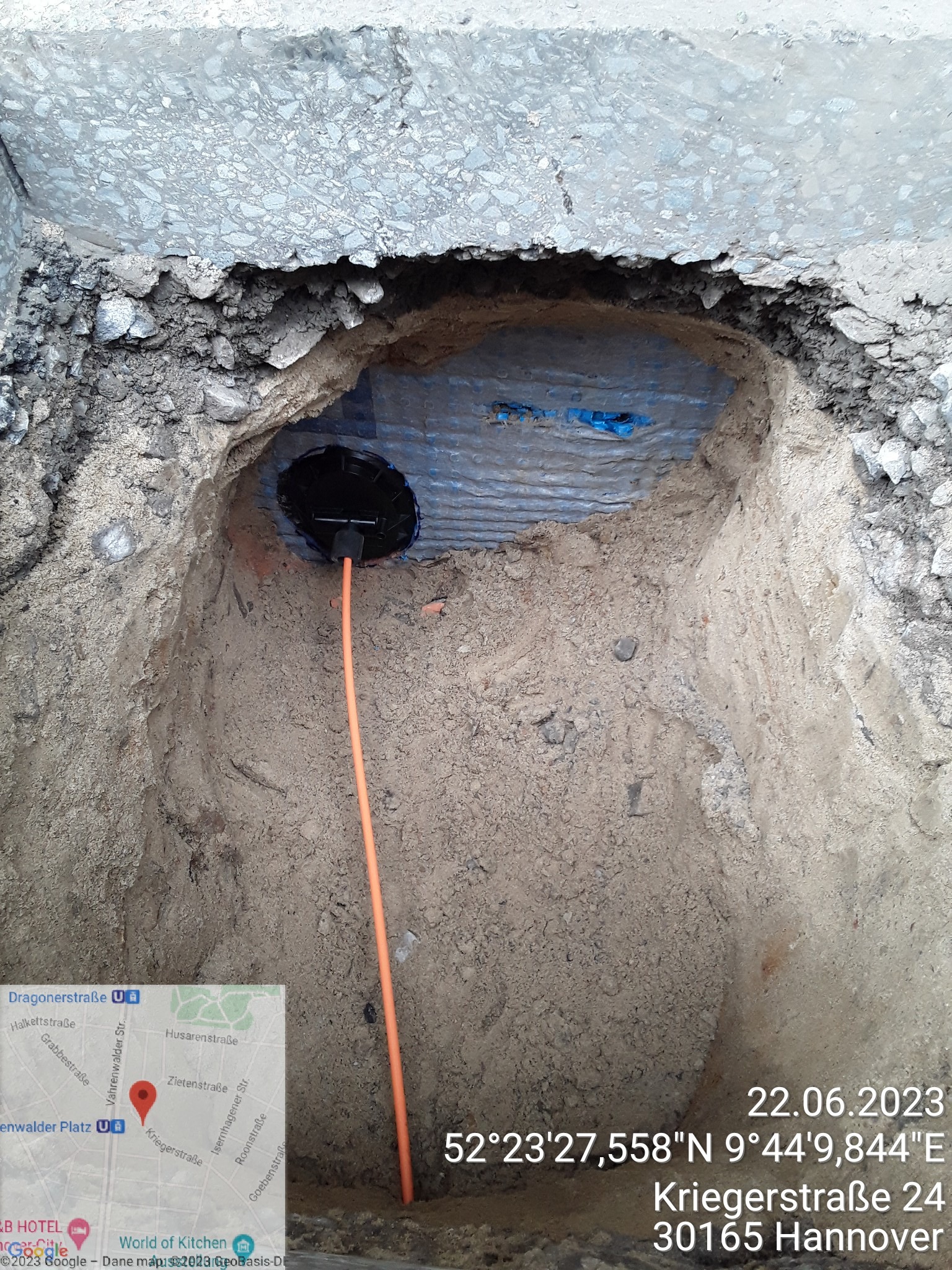 Wykop w ziemi z widoczną rurą osłonową i pomarańczowym przewodem, zabezpieczony niebieską folią, położony w pobliżu betonowej nawierzchni.