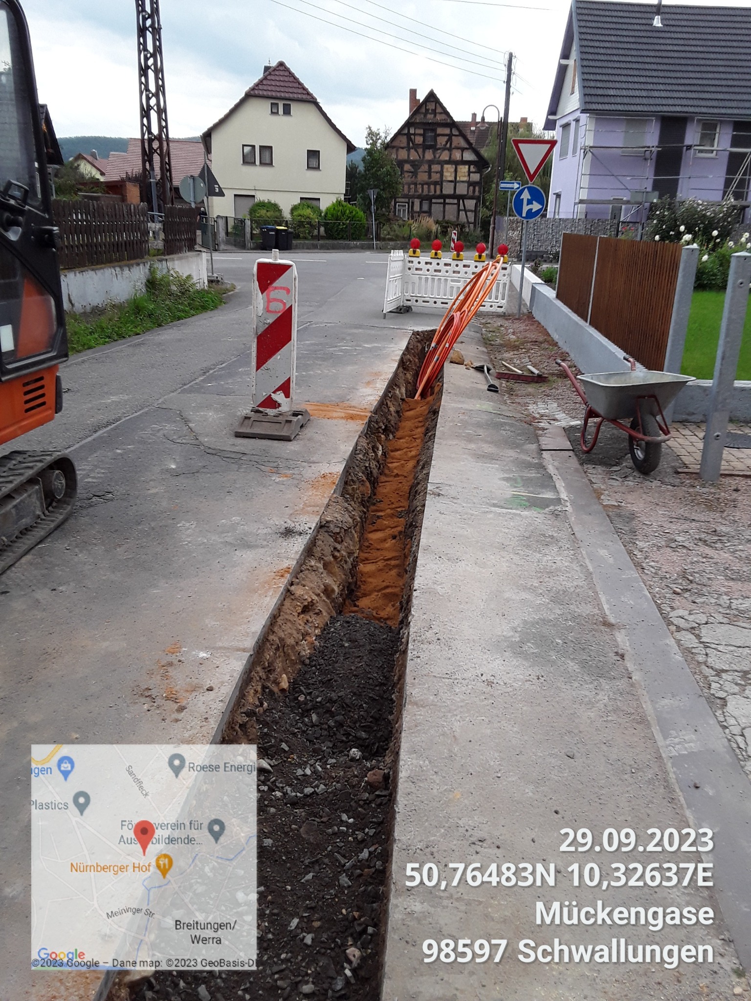 Wykopany wąski rów wzdłuż ulicy w małym miasteczku, pomarańczowe rury ułożone obok wykopu, widoczny mały traktor i taczka, tymczasowe bariery i znaki drogowe zabezpieczają teren.