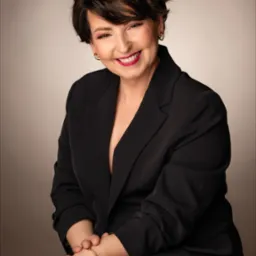 Portret uśmiechniętej kobiety w czarnej marynarce, z krótkimi, ciemnymi włosami i wyrazistym makijażem, pozującej na neutralnym tle. Złota bransoletka na nadgarstku dodaje elegancji.